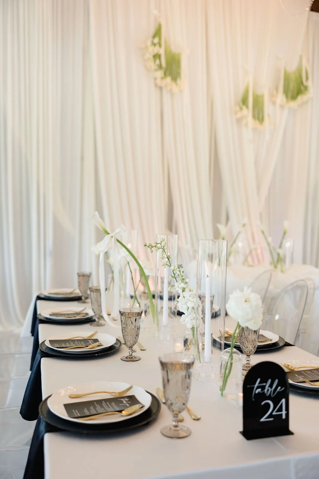 Elegant table setup with white and black dinnerware, gold utensils, tall glass candle holders, and white floral centerpiece, with a white curtain backdrop and a black table number sign reading 'Table 24'.