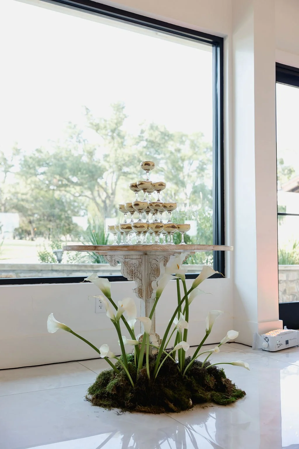 A tower of champagne glasses stacked in a pyramid shape on a wooden stand. The stand is decorated with white flowers and moss at the base. Large windows in the background show trees and outdoor scenery.