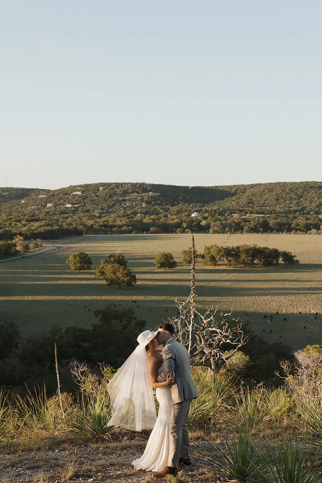 A bride and groom sharing a kiss outdoors during sunset, with a scenic landscape of open fields, trees, and rolling hills in the background.