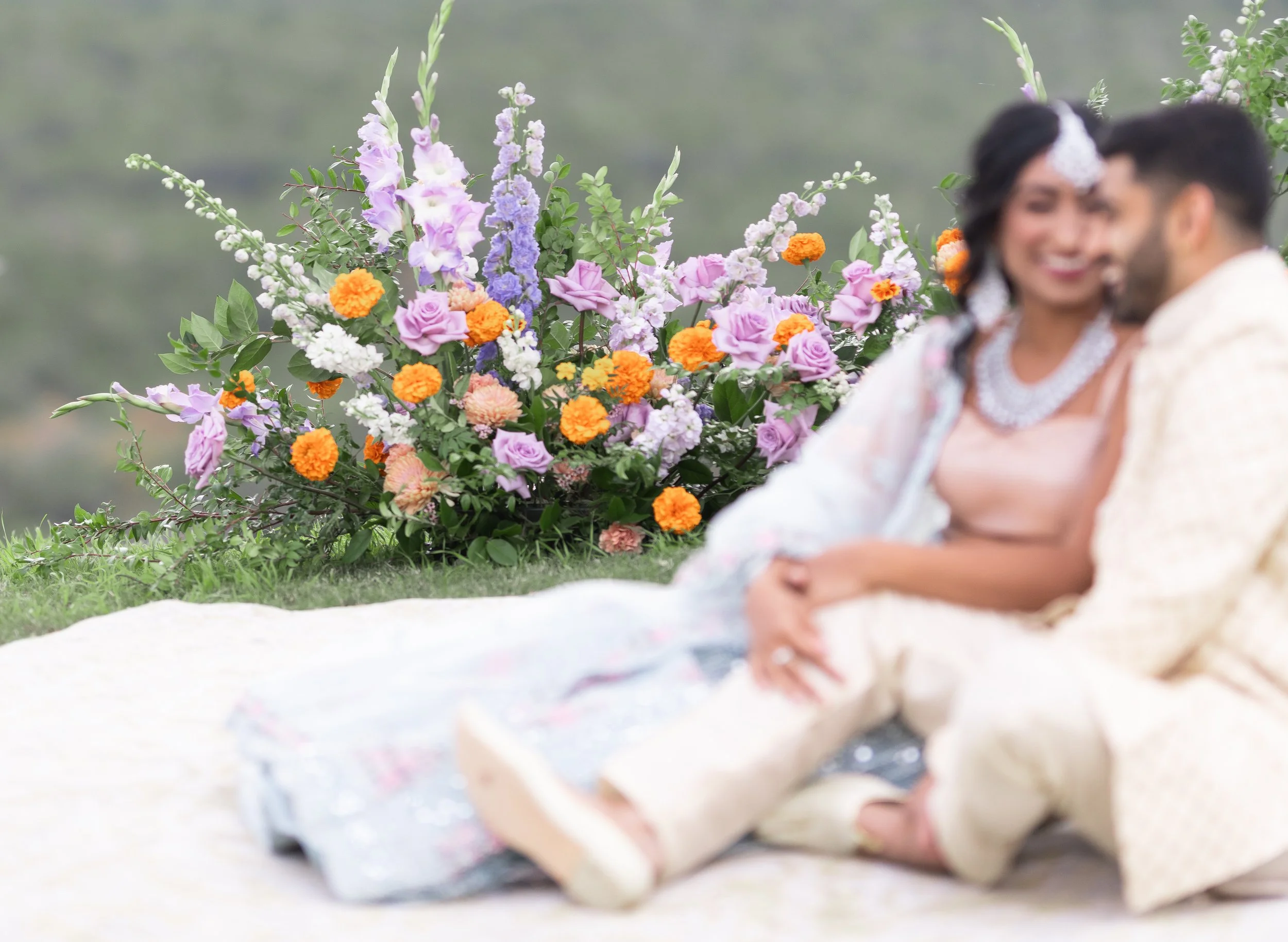 A newlywed couple sitting outdoors on a white rug, with a large, colorful flower arrangement in the background. The bride is wearing a pearl necklace and traditional wedding attire, and the groom is dressed in a cream-colored outfit. The focus is on 