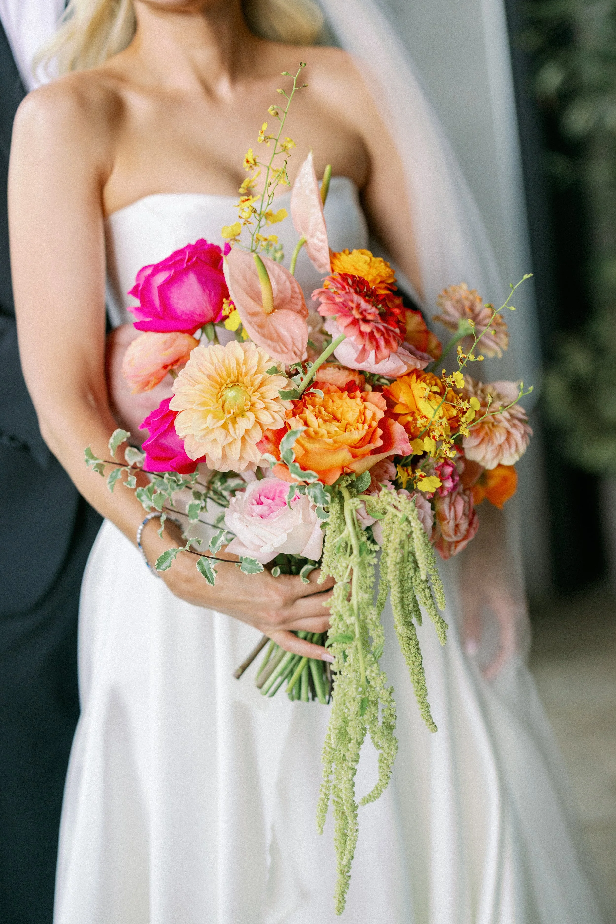 A bride in a strapless white wedding gown holding a large bouquet of colorful flowers, including pink, peach, yellow, and orange blooms with green foliage, at her wedding.