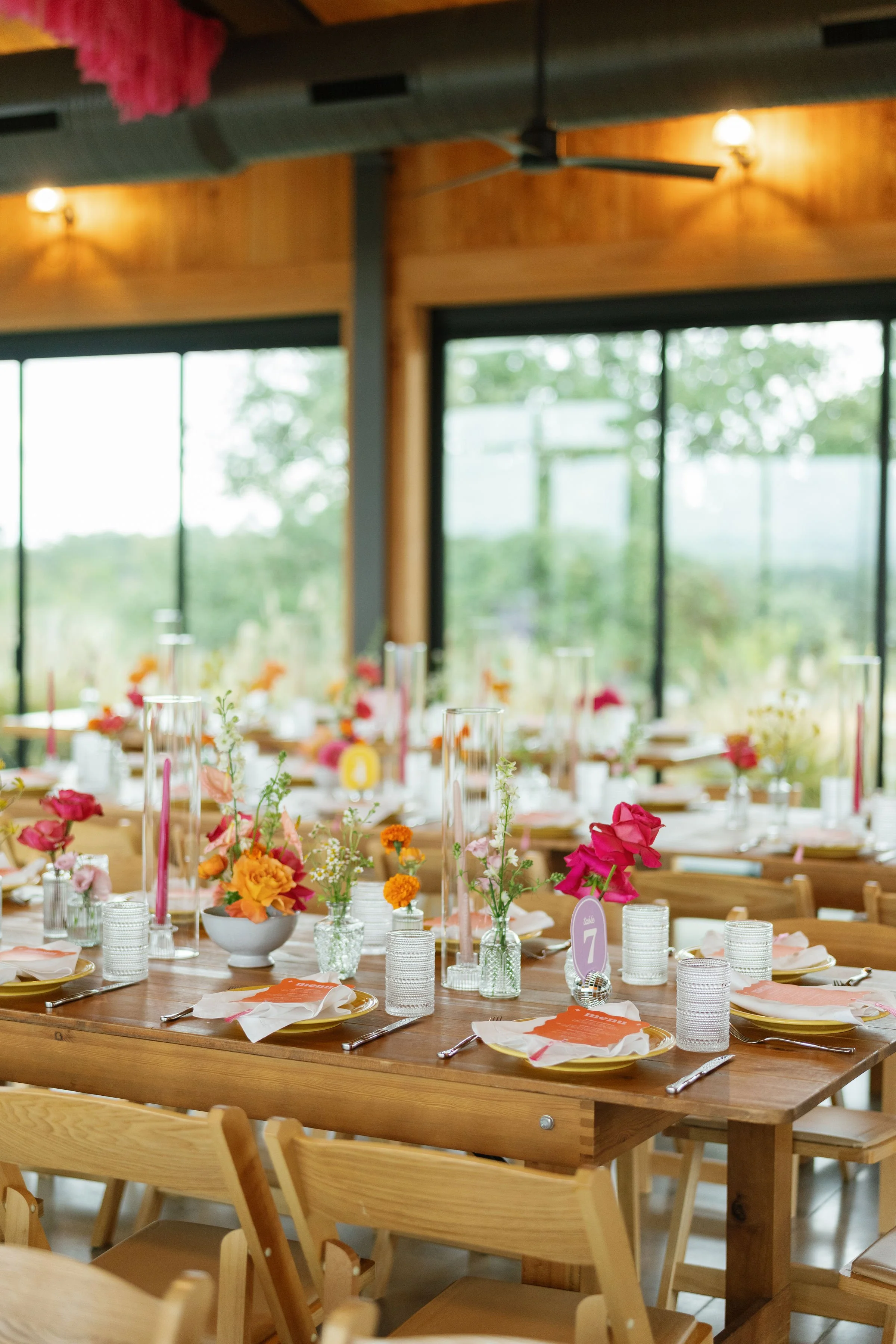 A beautifully decorated dining table with colorful flowers, candles, and tableware in a bright, airy room with large windows and wooden accents.