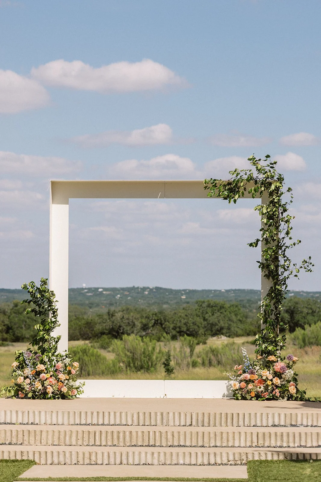 A white wedding arch decorated with greenery and pink and white flowers, set outdoors against a landscape of greenery and a partly cloudy sky.