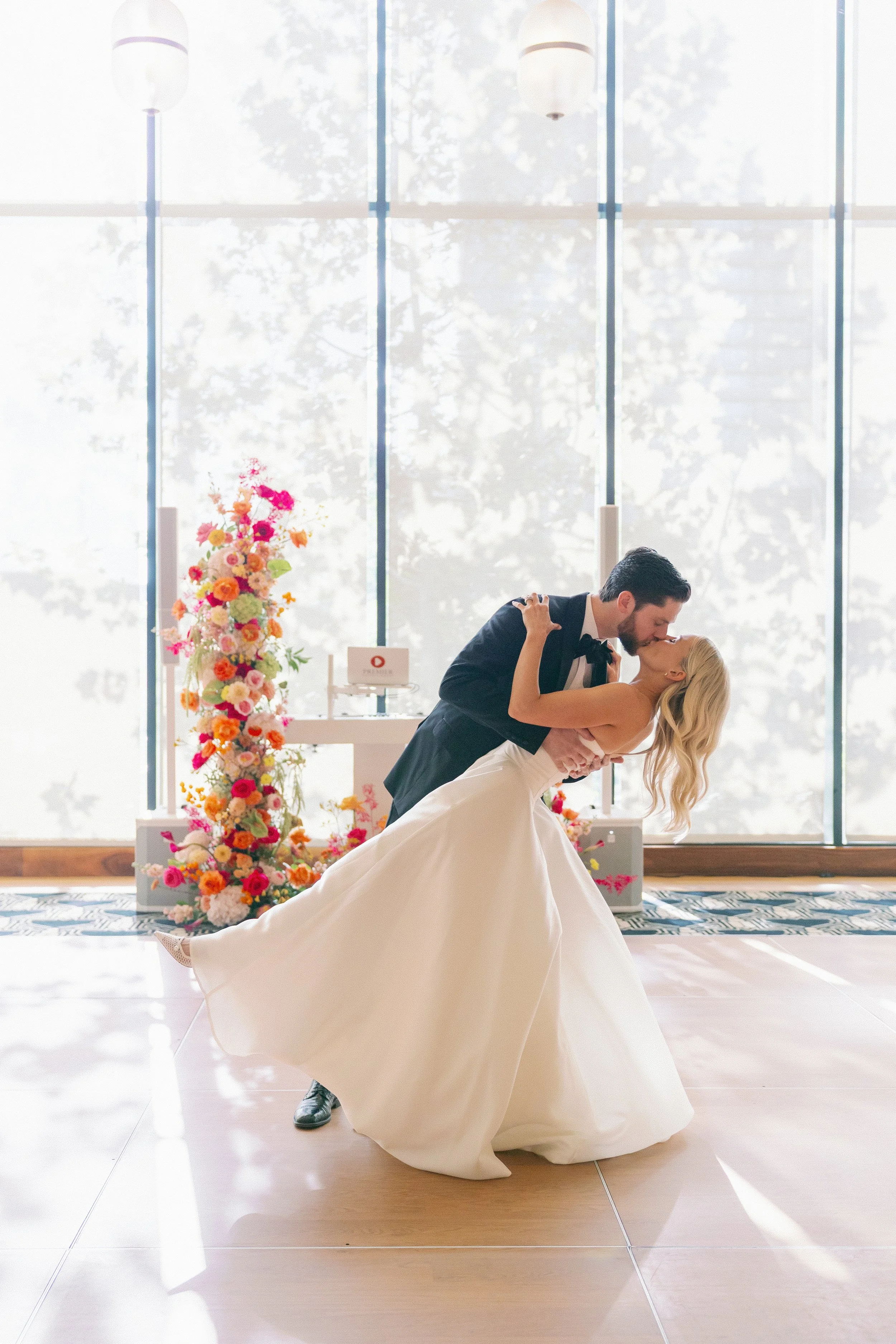 A bride and groom sharing a kiss during their wedding ceremony, with a floral arrangement and large windows in the background.