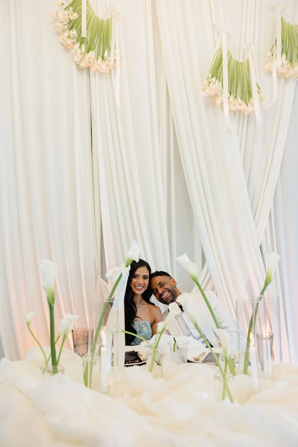 A couple sitting at a wedding reception table, smiling, with white flowers and pink floral arrangements behind them and white drapery as a backdrop.