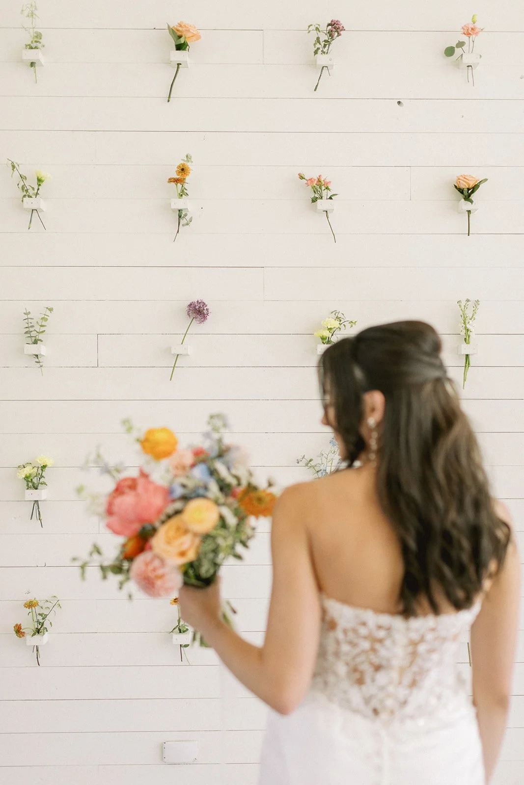 A woman in a lace strapless dress with long wavy hair holding a colorful bouquet of flowers, standing in front of a white wall decorated with small vases of various colored flowers.