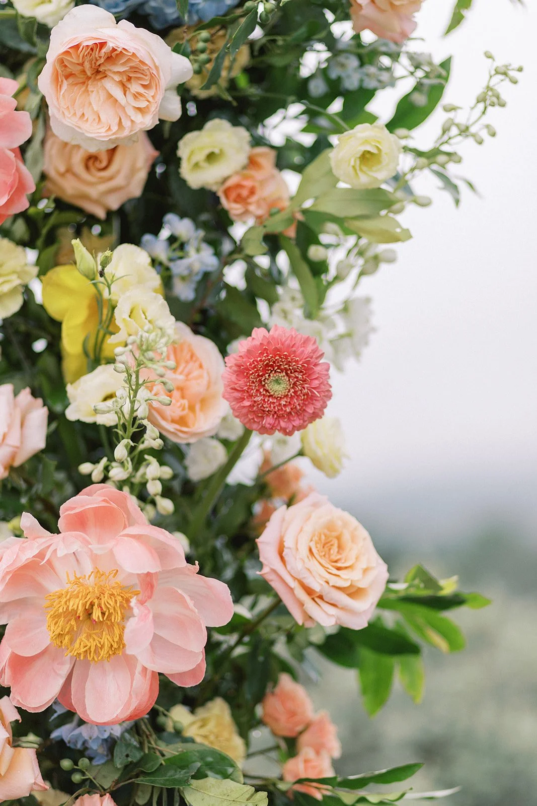 Colorful bouquet of various flowers including roses, a pink gerbera daisy, and other blossoms against a blurred background.