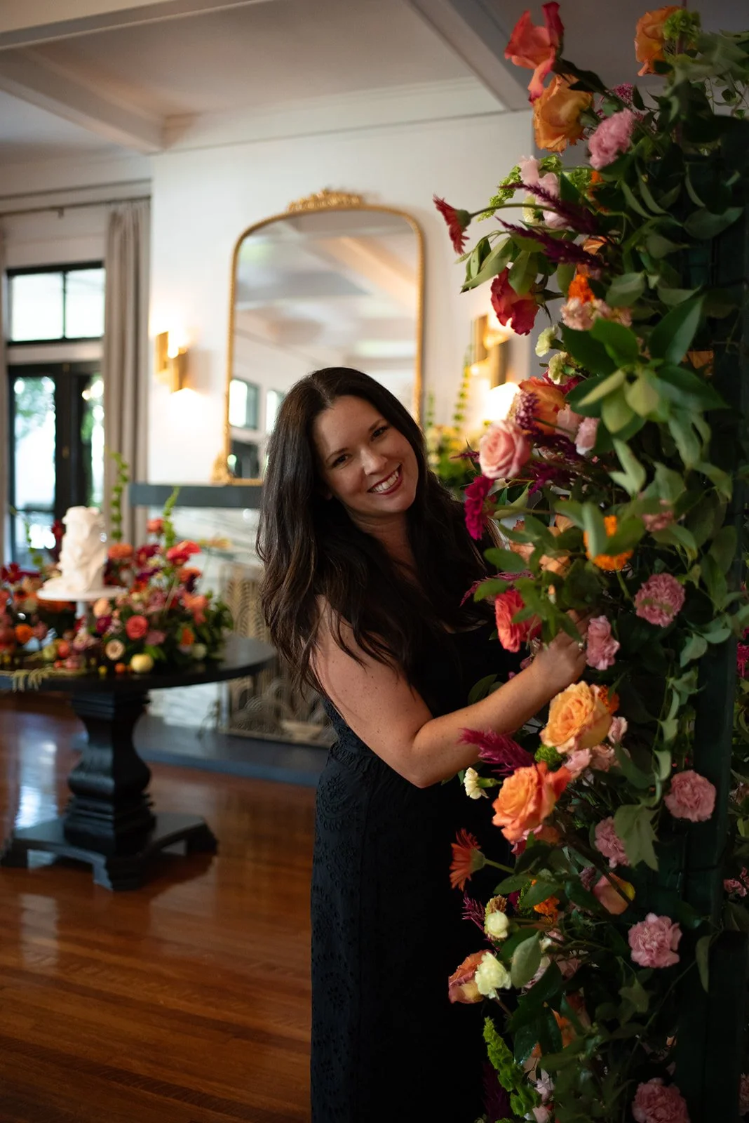 Woman with long dark hair smiling at the camera, partially hidden behind a floral arrangement with pink, orange, and red flowers in an elegant indoor setting.
