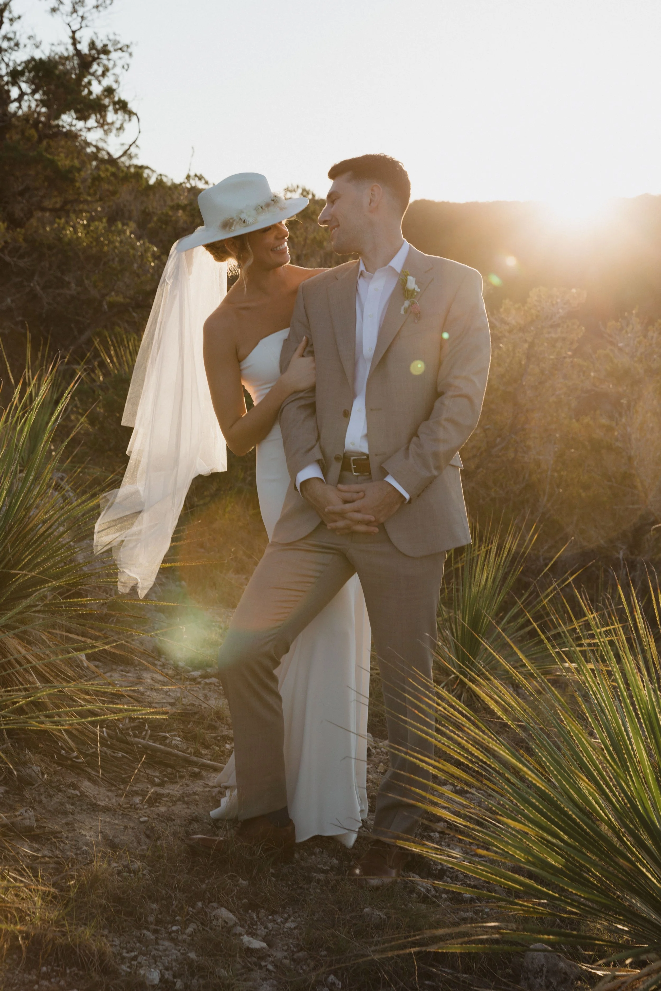 A bride and groom standing outdoors at sunset, with the bride wearing a white dress and wide-brimmed hat, and the groom in a beige suit, smiling and looking at each other amidst desert vegetation.