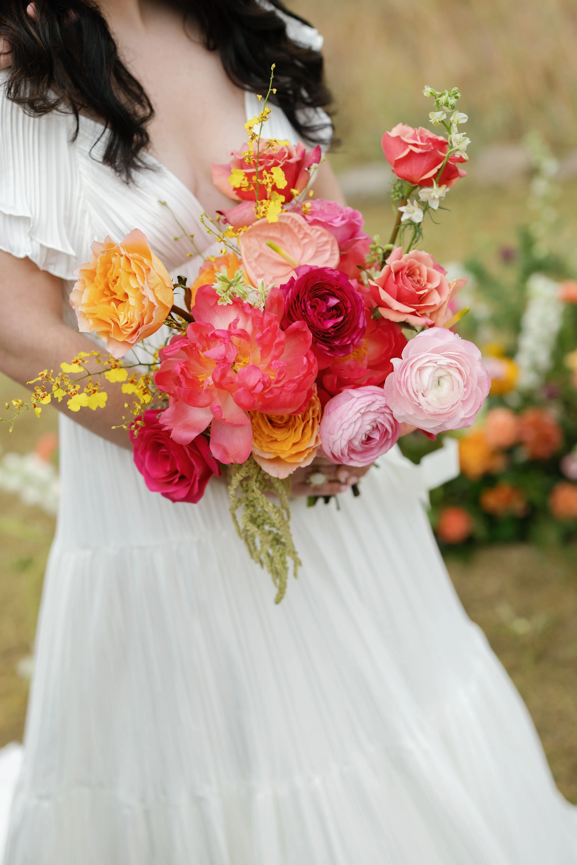Woman in white dress holding a colorful bouquet of pink, orange, red, and yellow flowers in an outdoor setting.