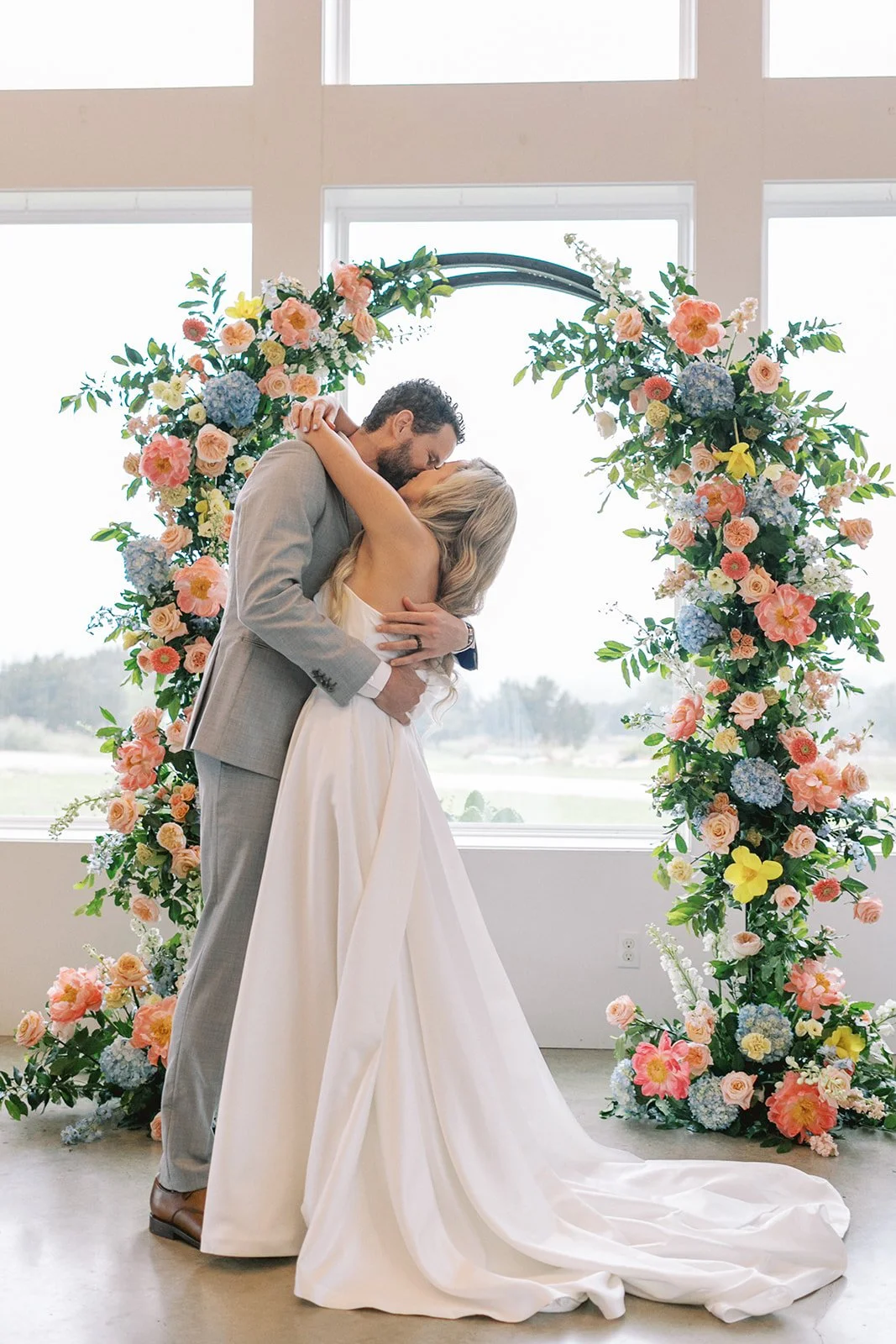 A couple kissing during their wedding ceremony in front of a floral arch with pink, blue, and cream flowers, and large windows in the background.