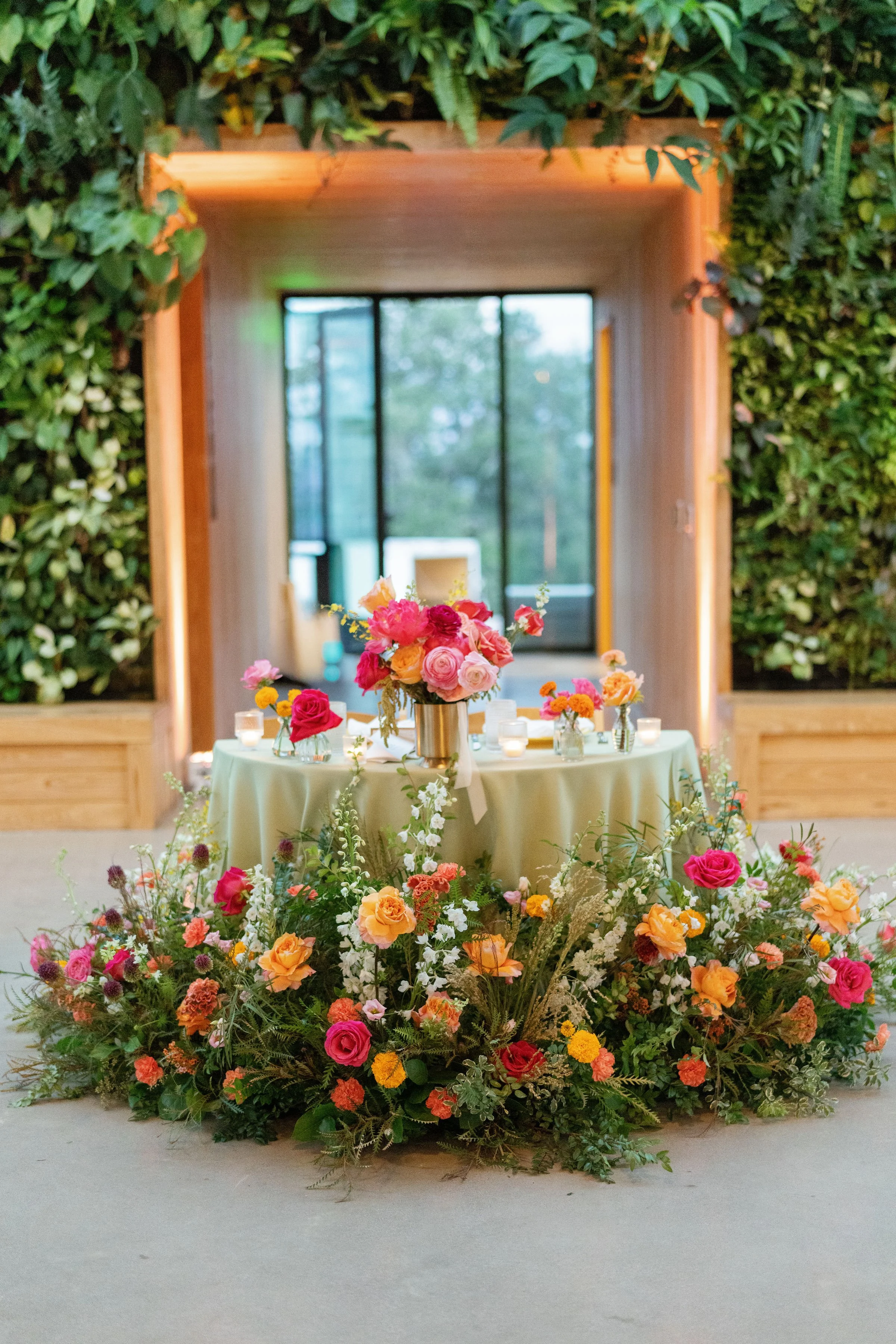 A table decorated with a floral arrangement of pink, orange, and yellow flowers, surrounded by the same type of flowers and greenery at the base, set in a room with a wooden arch and a large window in the background.