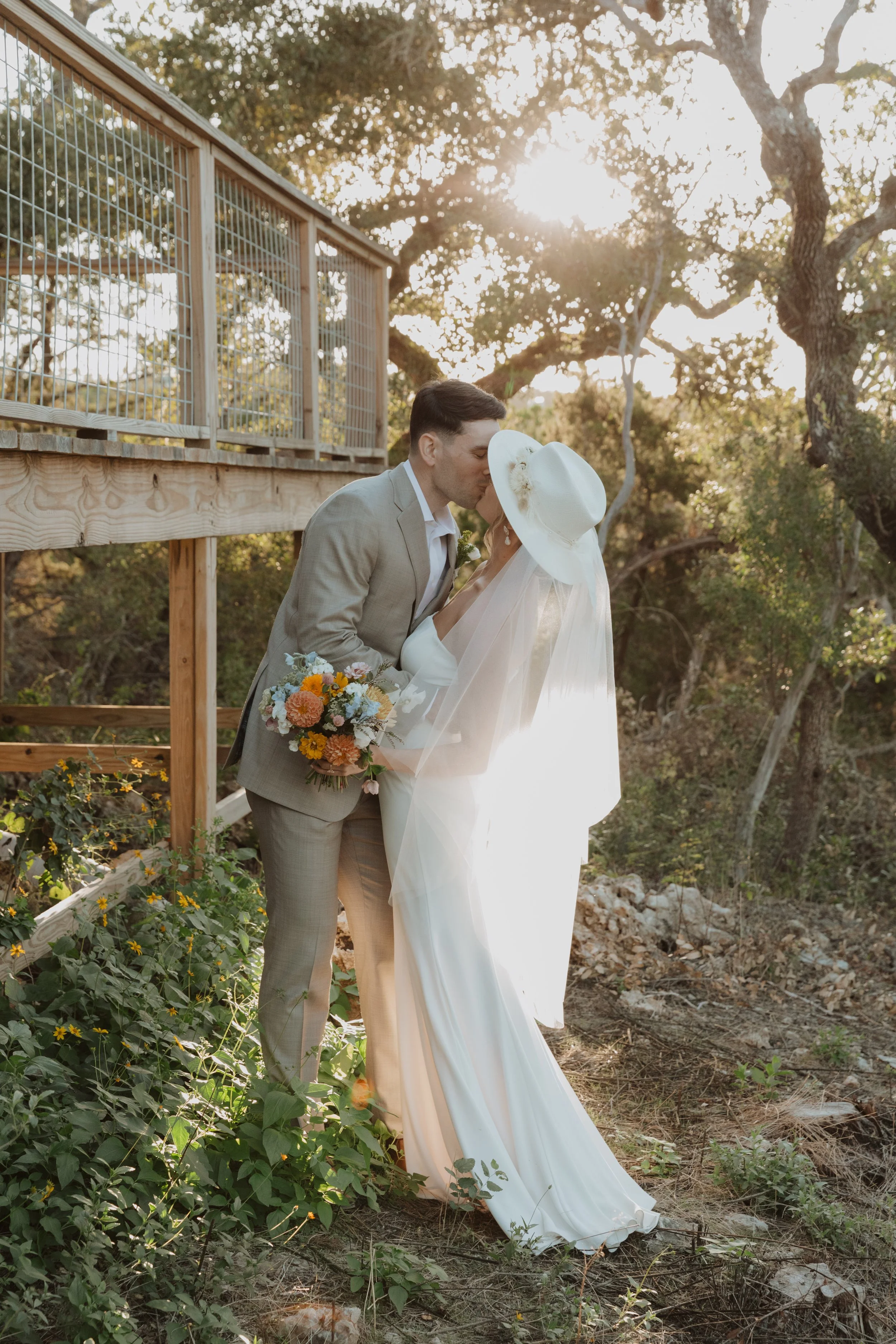A couple dressed in wedding attire sharing a kiss outdoors during sunset, with trees and foliage in the background.