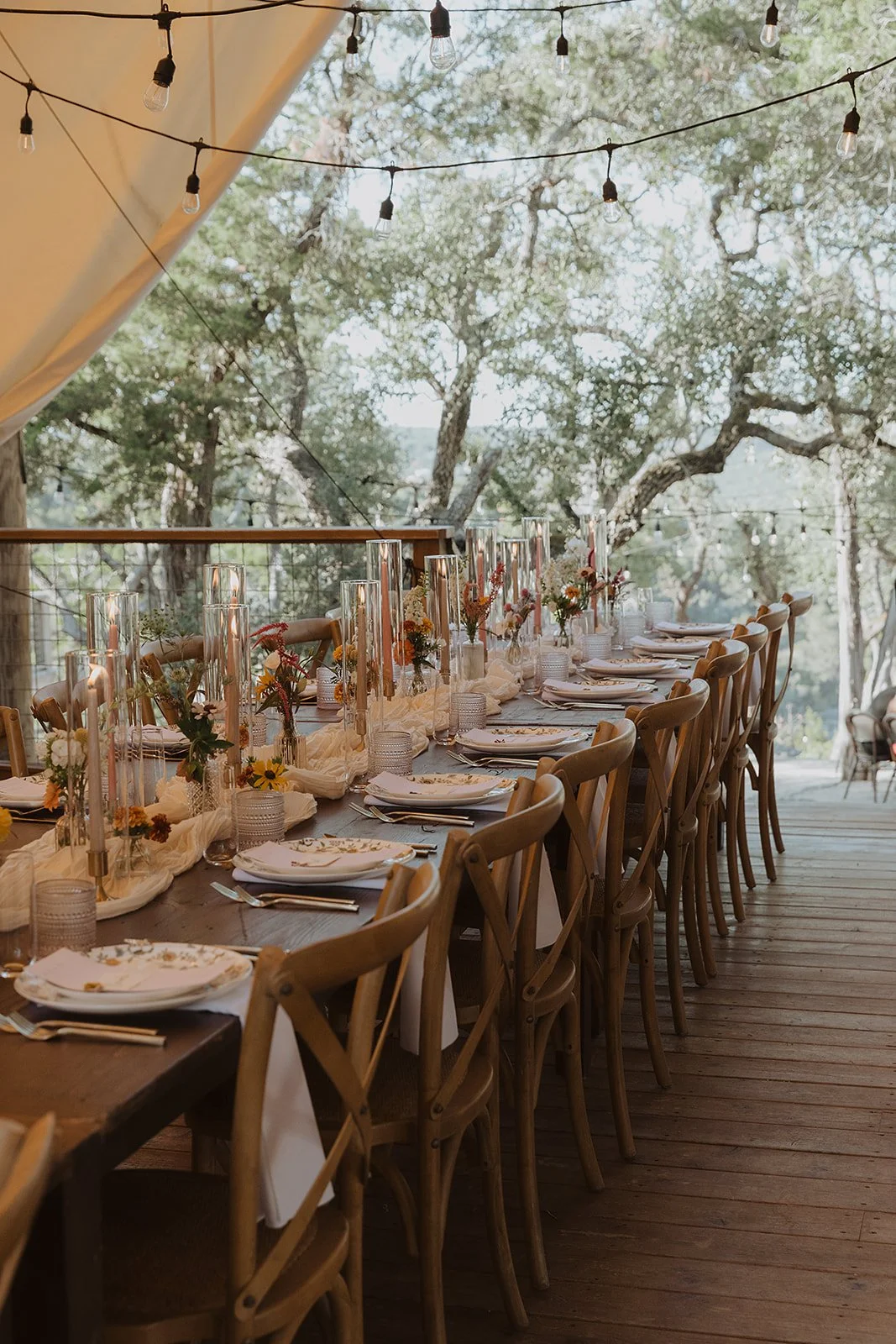 Long wooden dining table set with white plates, silverware, and glasses, decorated with floral centerpieces and candles, outdoors with string lights and trees in the background.