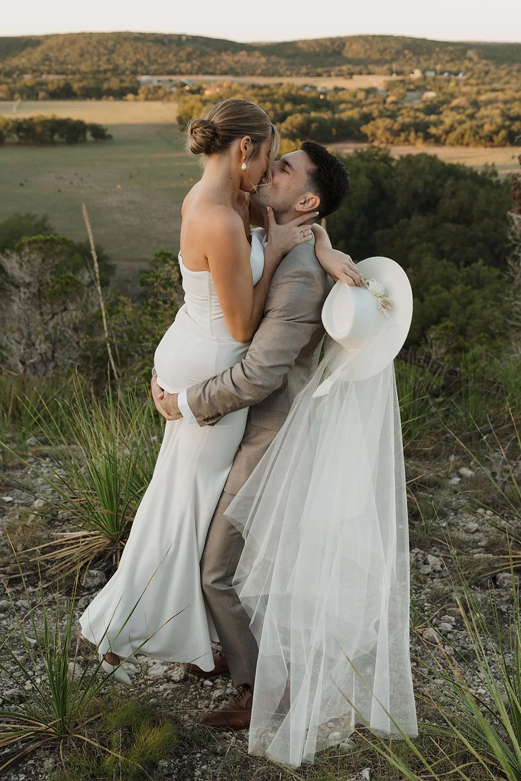 A couple in wedding attire sharing an embrace outdoors, with a scenic landscape of hills and trees in the background.