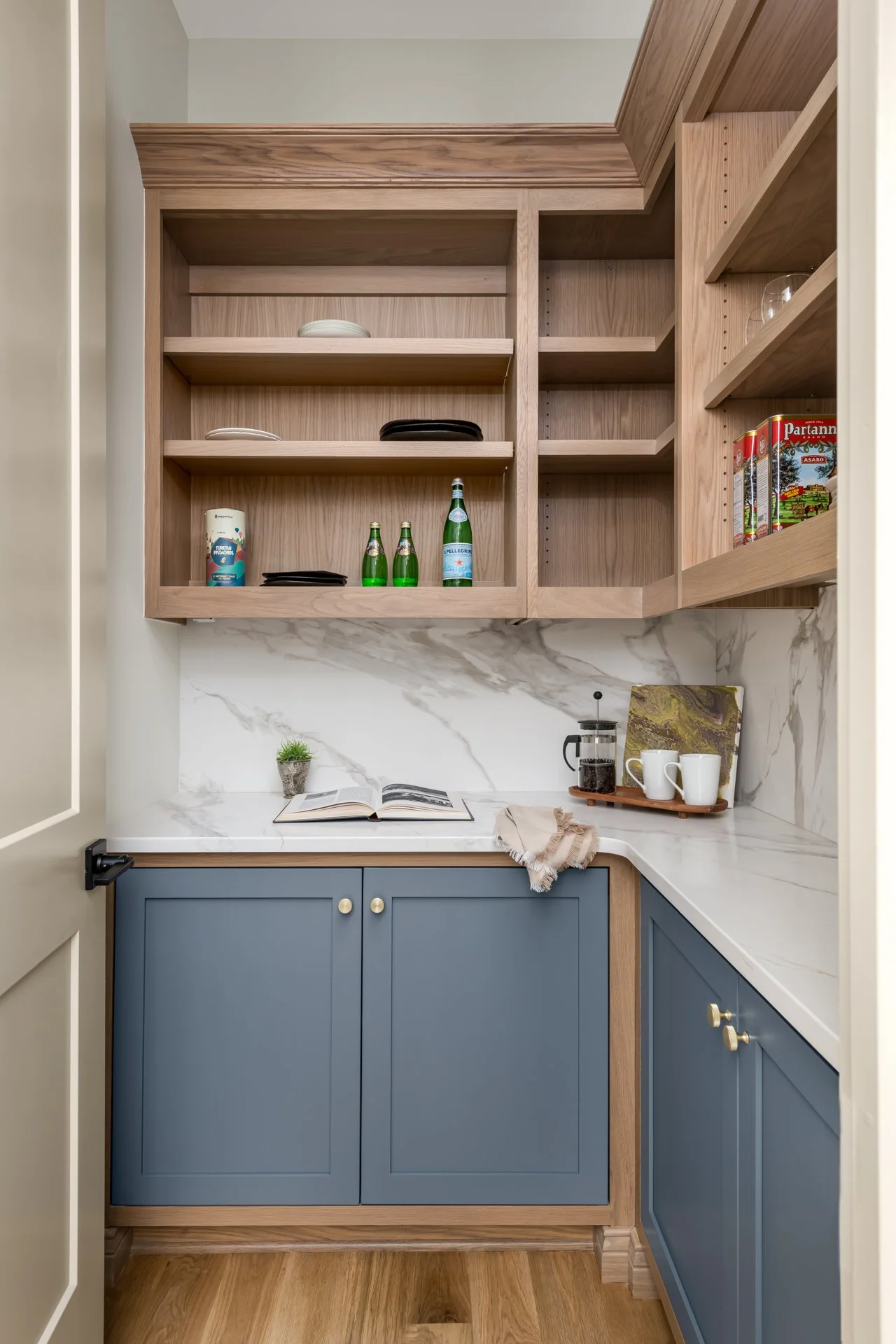 Tucked behind the main kitchen, this functional scullery features custom blue cabinetry with brushed gold hardware, natural wood open shelving, and a marble-look quartz countertop and backsplash. Designed for hidden prep and storage, it's a chef’s dr