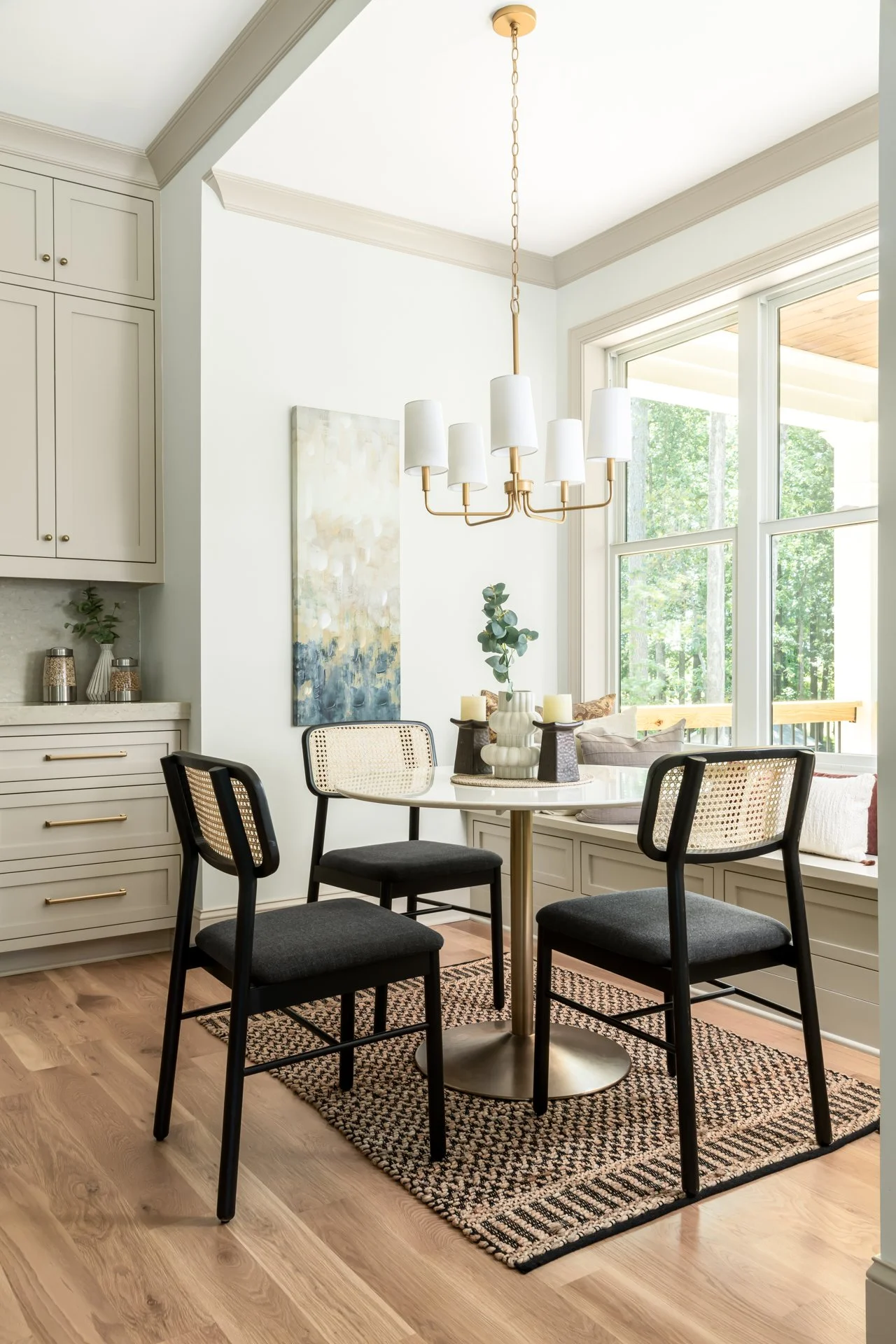 This cozy breakfast nook offers a sun-drenched retreat with built-in bench seating, a round marble pedestal table, and woven-back dining chairs. Surrounded by tall windows and styled with warm neutral cabinetry and a sleek gold chandelier, it’s the p