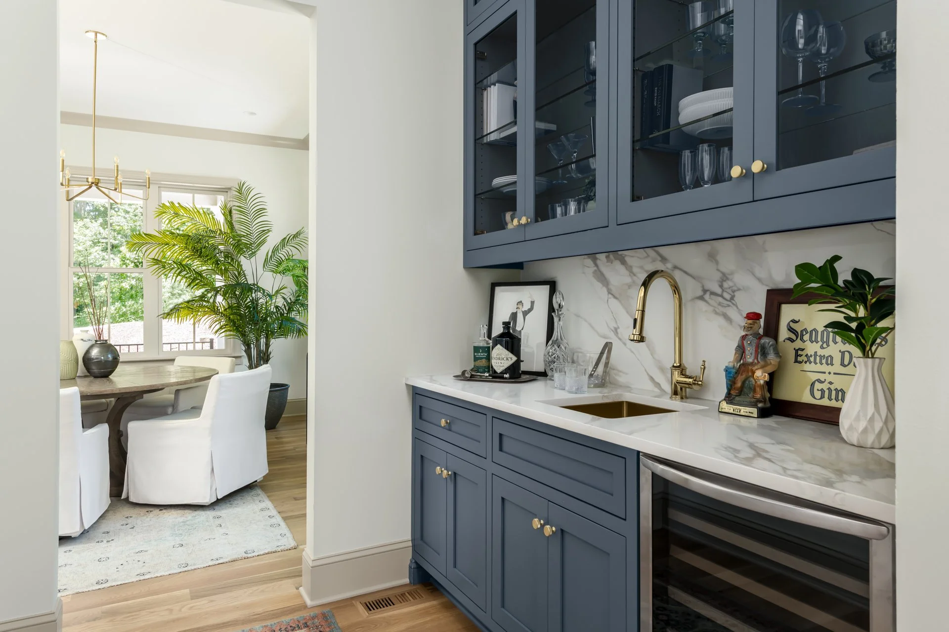 This striking wet bar features deep navy blue cabinetry, marble-look quartz countertops, and glass display cabinets. A gold faucet and bar sink add a luxe feel, while integrated wine storage and decor-ready shelving make this the perfect space to ent