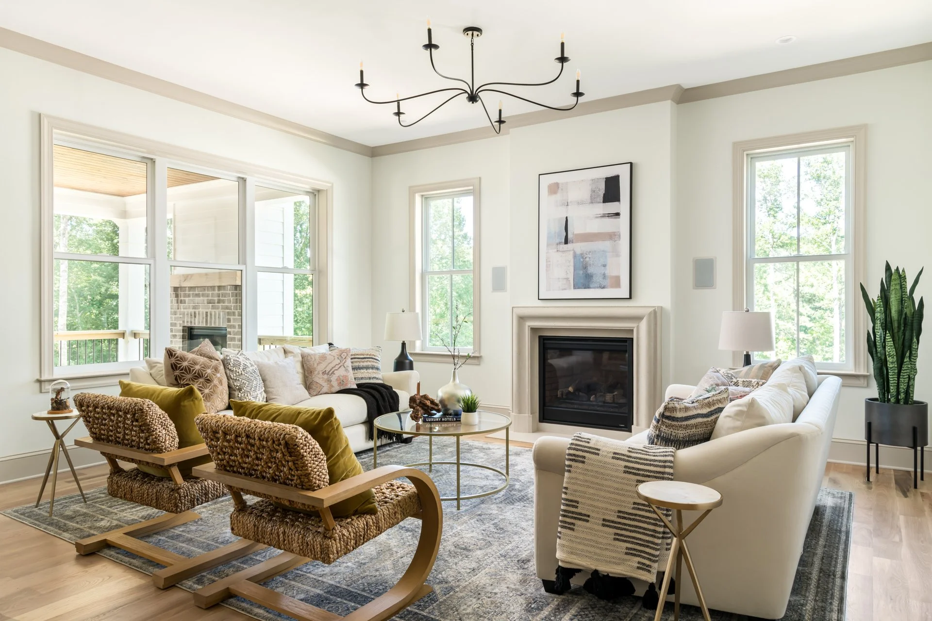 The living room showcases a serene color palette, oversized windows for ample natural light, and a sleek gas fireplace with a cast stone surround. Curated modern furniture, a statement chandelier, and woven accent chairs bring texture and personality