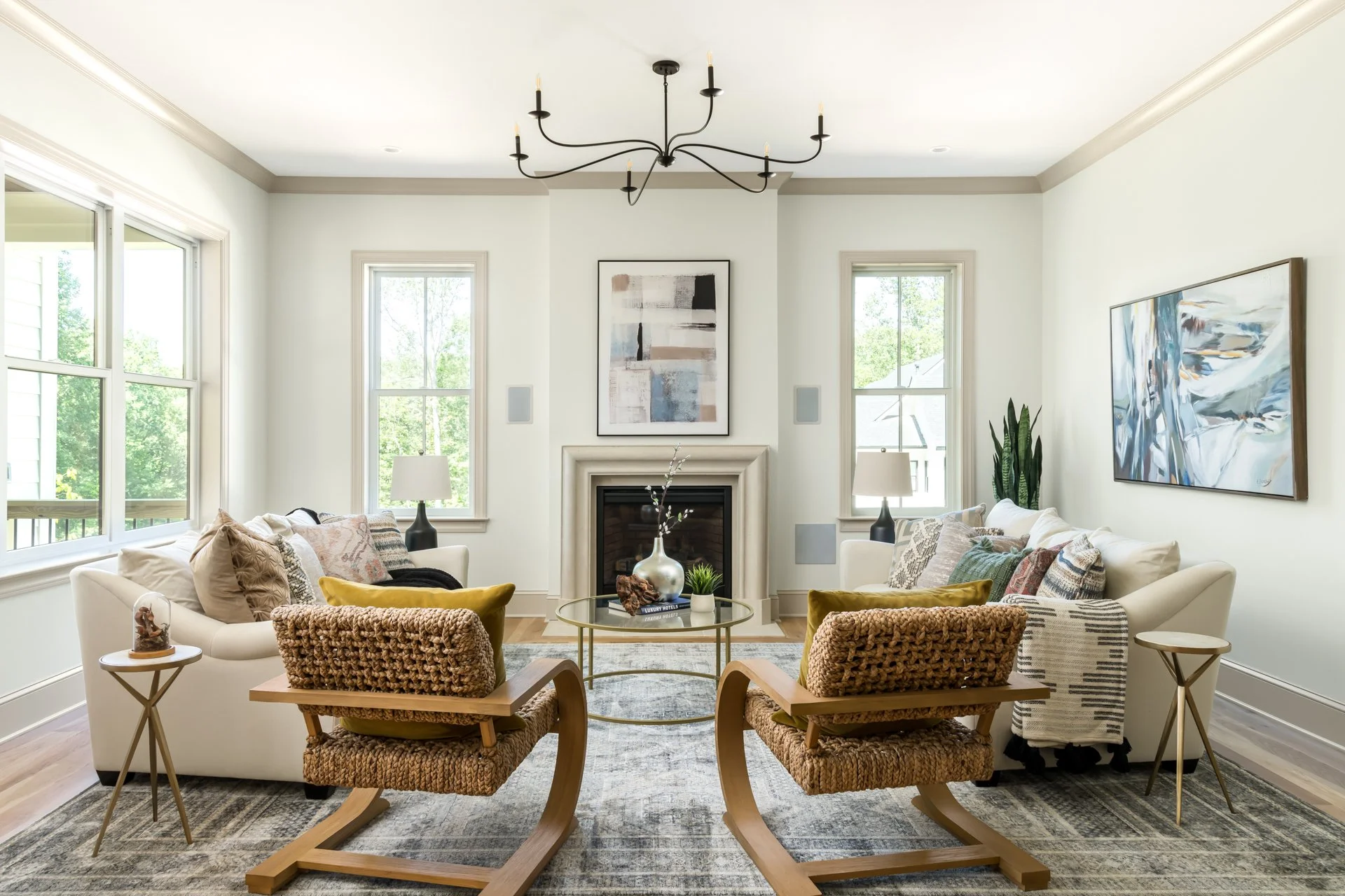 The living room showcases a serene color palette, oversized windows for ample natural light, and a sleek gas fireplace with a cast stone surround. Curated modern furniture, a statement chandelier, and woven accent chairs bring texture and personality