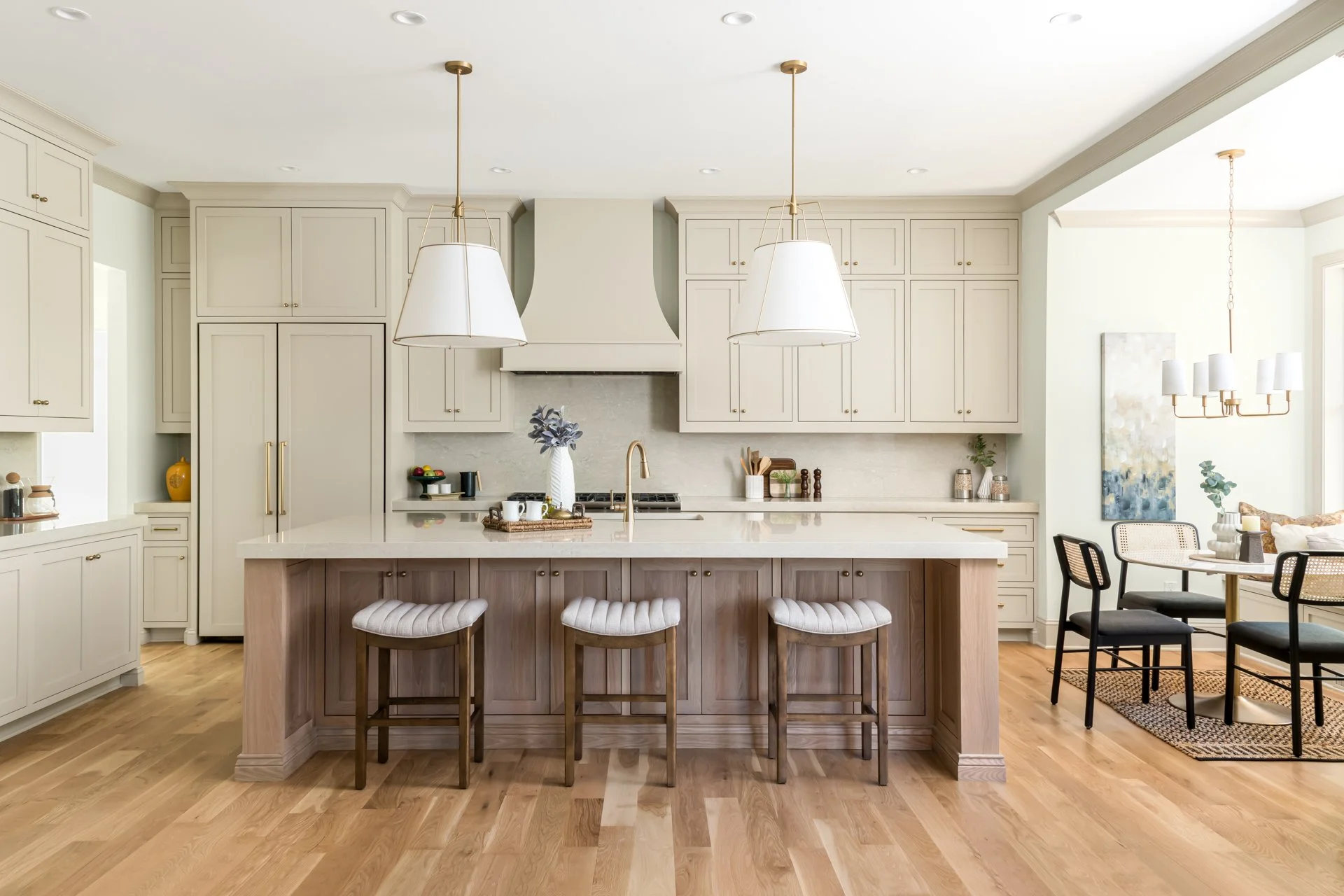 This elegant kitchen features floor-to-ceiling custom cabinetry in a warm neutral tone, a professional-grade gas range with custom hood, and an oversized quartz island perfect for entertaining. Brass fixtures, pendant lighting, and white oak wood flo