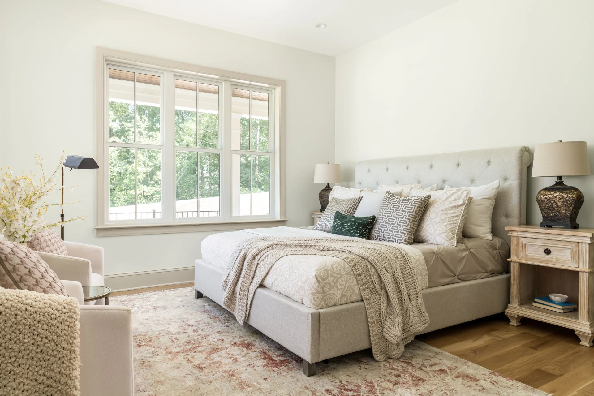 Designed for rest and relaxation, the primary bedroom features an upholstered tufted bed, soft neutral bedding, layered pillows, and natural wood nightstands. Large windows allow soft morning light to flood in, and a pair of blush armchairs with a fl