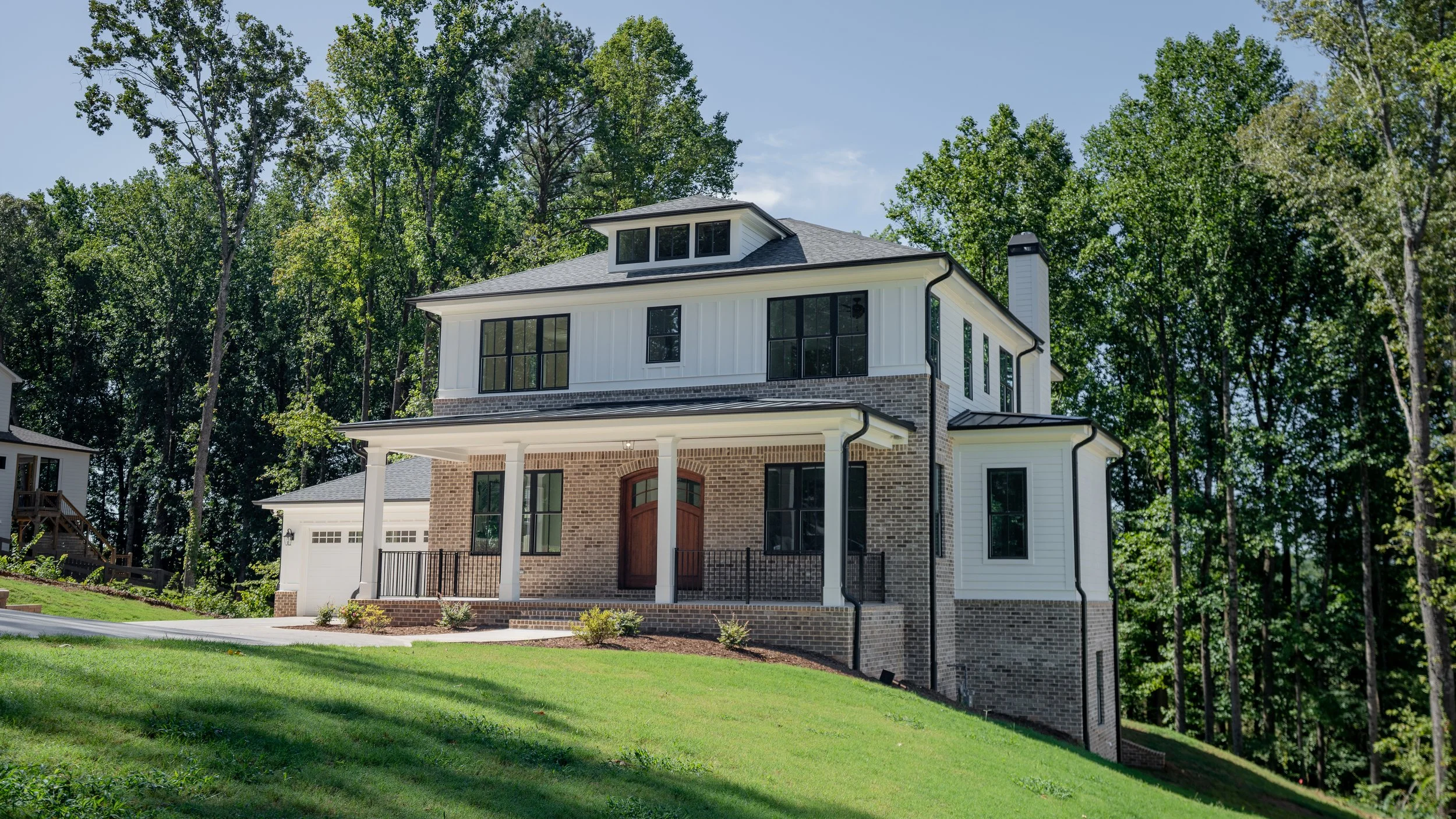Modern multi-story house with brick and white siding exterior, featuring large black-framed windows, a covered front porch with columns, and surrounded by a well-maintained lawn and tall trees.