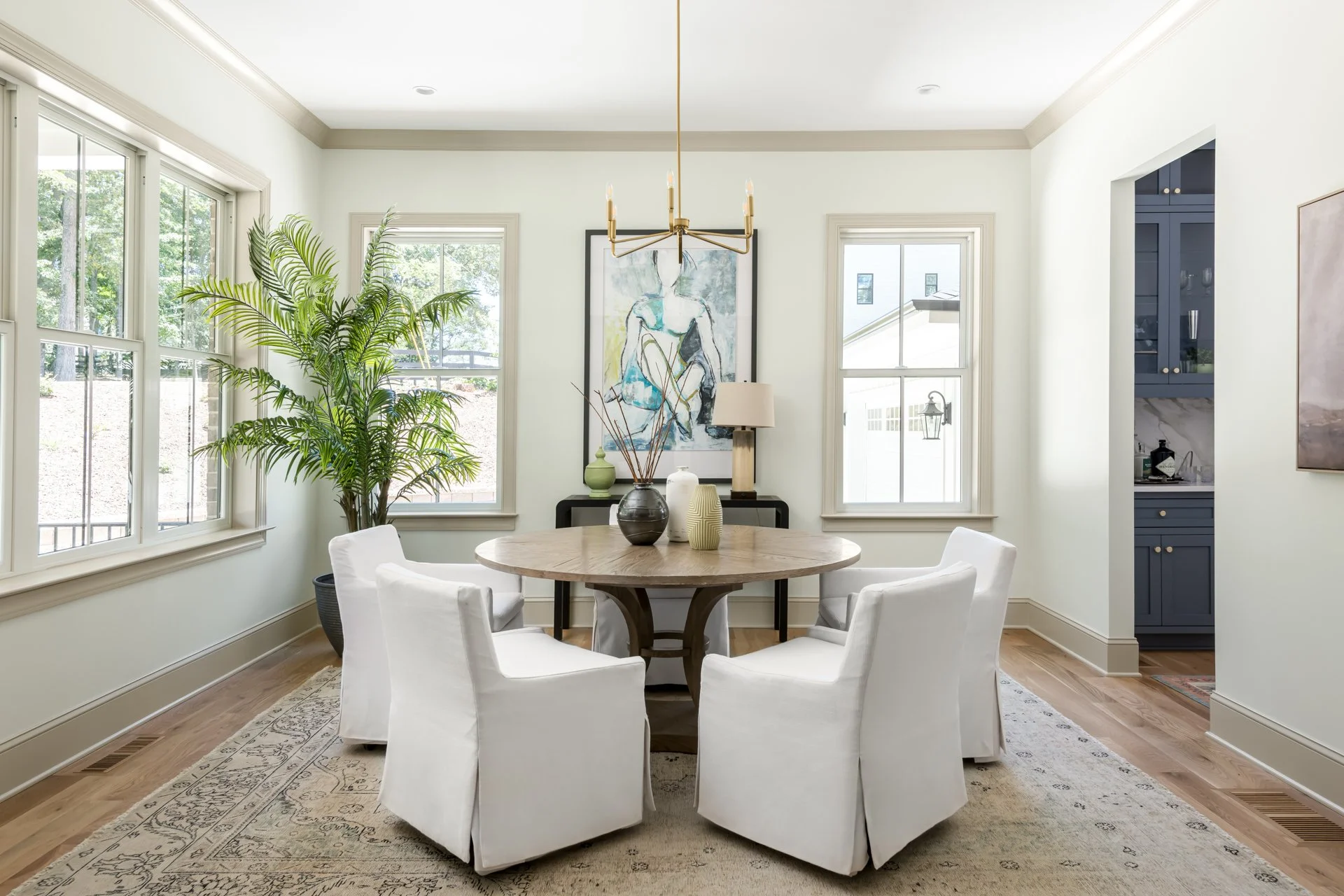 Flooded with natural light, the dining room features a round wood pedestal table, tailored white slipcovered chairs, and modern chandelier lighting. Framed art and a decorative console table round out this polished yet inviting dining space with view