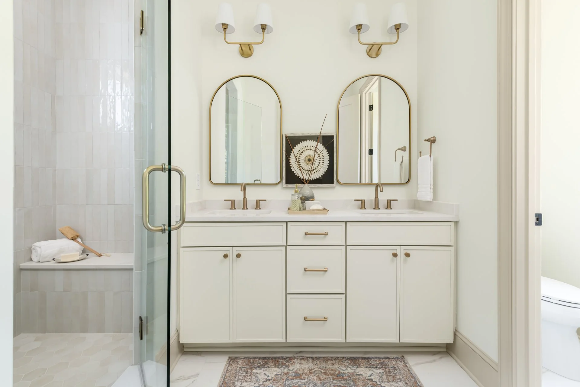 This refined bathroom features dual arched mirrors above a double-sink vanity with soft-close cabinetry and brushed gold fixtures. The walk-in shower includes handmade-look wall tiles, a built-in bench, and frameless glass for a clean, spa-like finis
