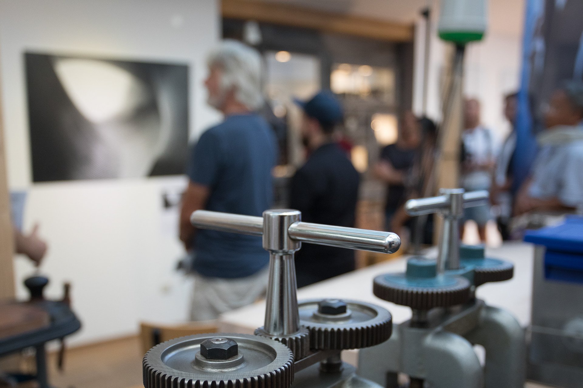 Nahaufnahme von Metallgetrieben im Vordergrund, mit unscharfen Menschen im Hintergrund, die in einem Raum eine Kunst- oder Fotografieausstellung betrachten.