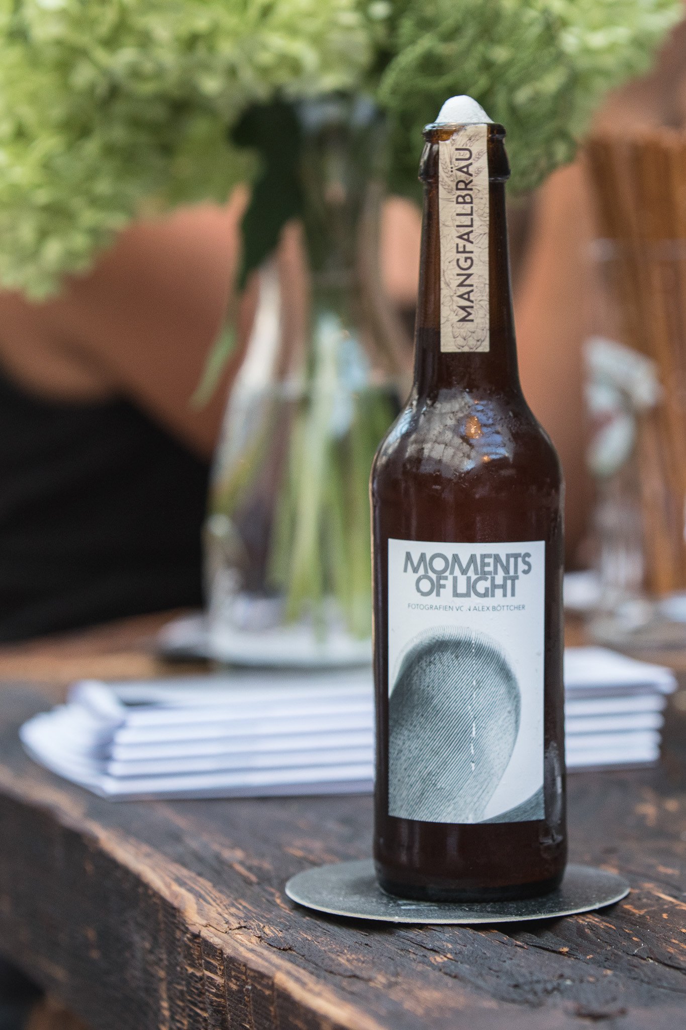 Eine Bierflasche "Moments of Light" von Mangfallbrau auf einem Holztisch, im Hintergrund Dekoration wie Blumen und Bücher.