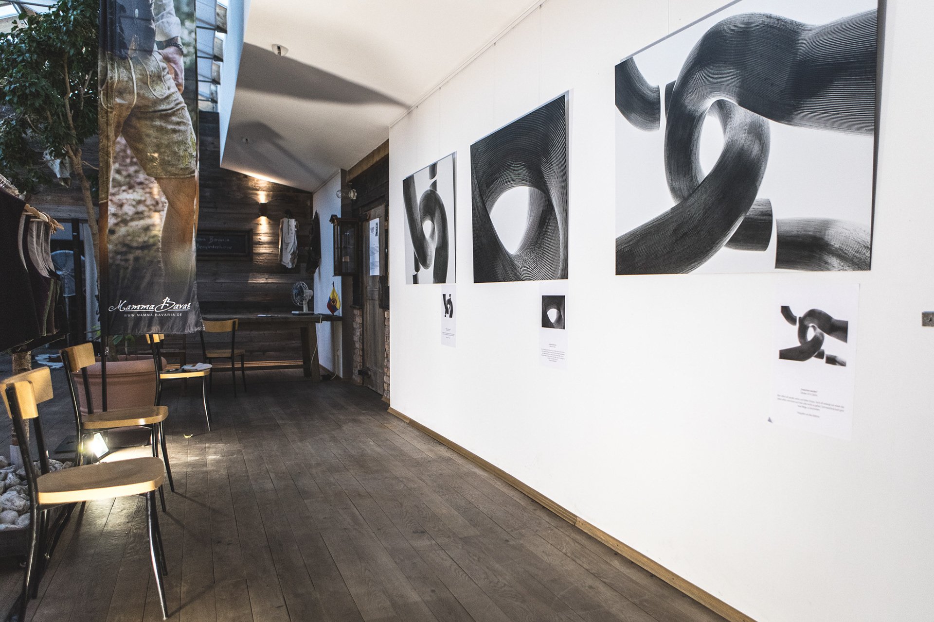 Galeriebereich mit modernen Kunstwerken an der Wand, Holzboden und Stühlen. Ein Banner mit Bild einer Person in Tracht hängt seitlich.