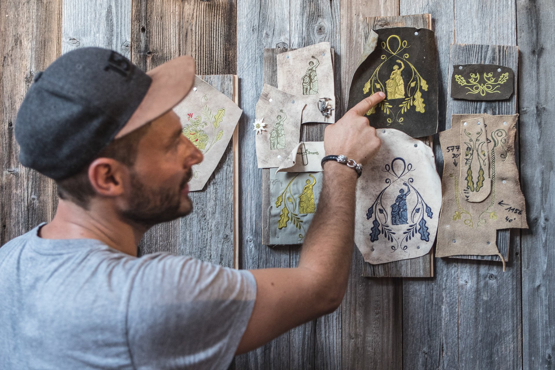 Mann zeigt auf handgemalte Designs auf Papierstücken, die an einer Holzwand befestigt sind.