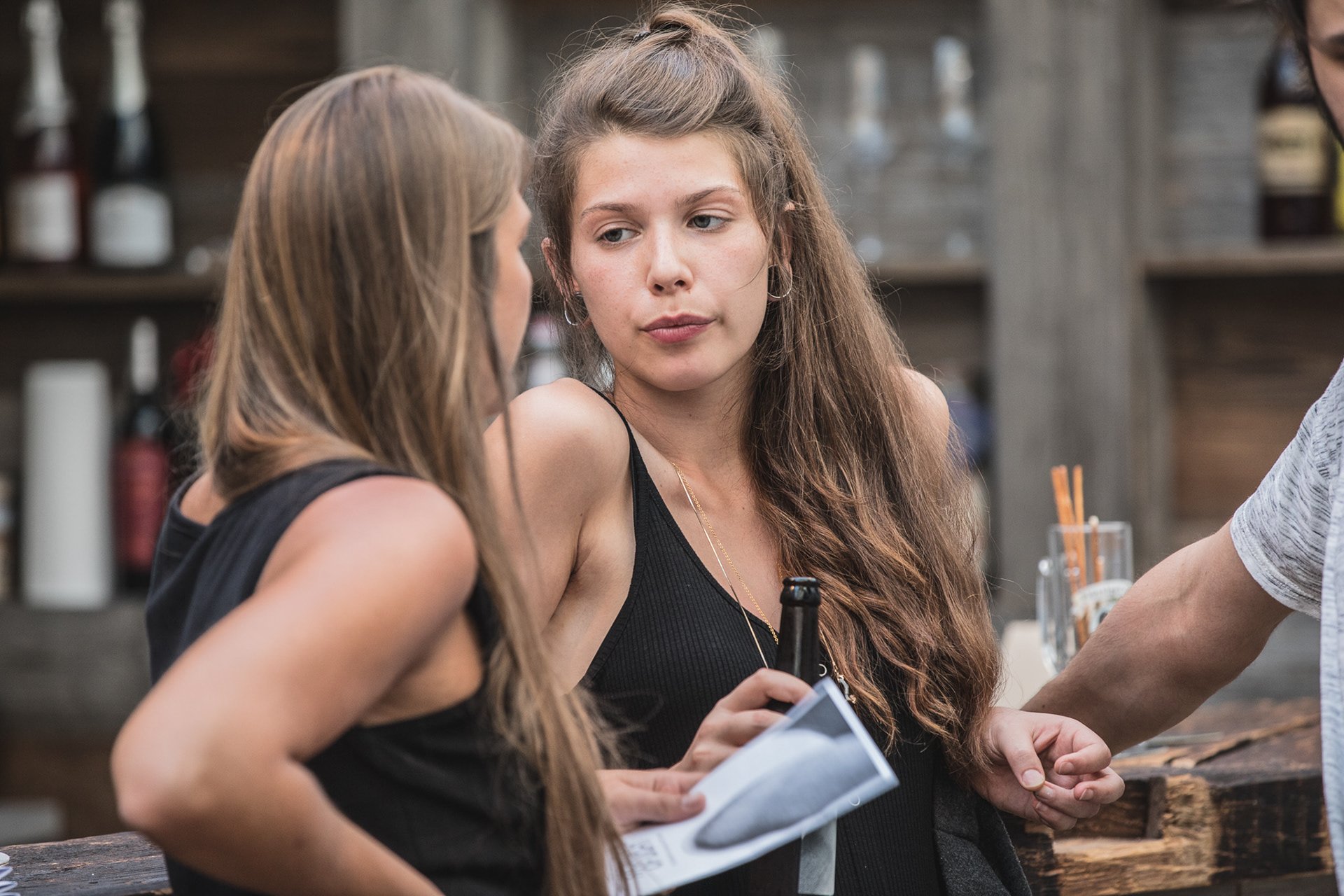 Zwei Frauen unterhalten sich in einer Bar. Die Frau links hält ein Blatt Papier, während die Frau rechts eine Bierflasche in der Hand hat. Im Hintergrund sind Flaschen und Gläser zu sehen.