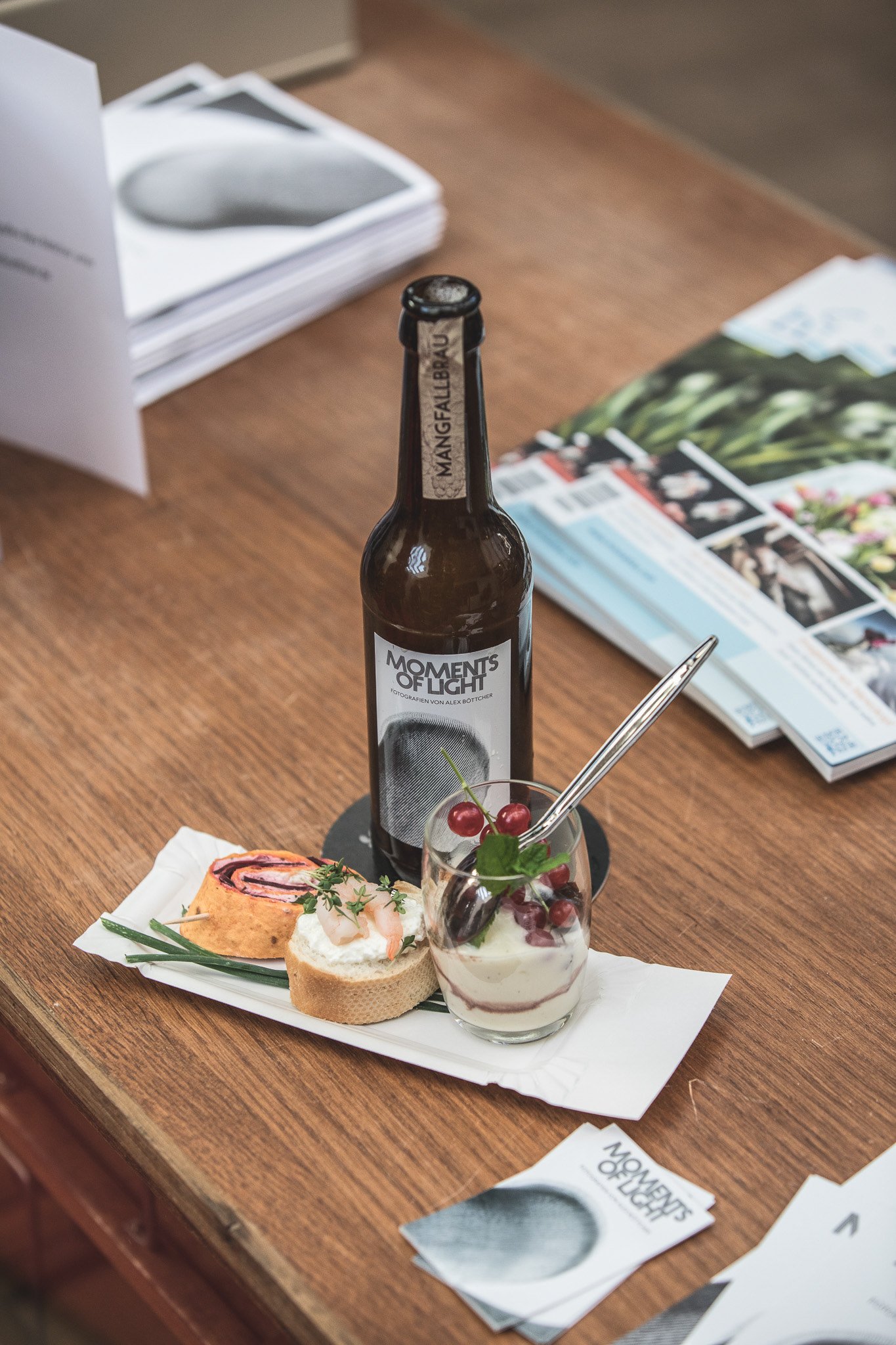 Holztisch mit einer Bierflasche "Moments of Light", einer kleinen Platte mit Gebäck und Garnelen, einem Dessertglas mit Sahne und Beeren. Im Hintergrund sind Broschüren und ein Papierstapel zu sehen.