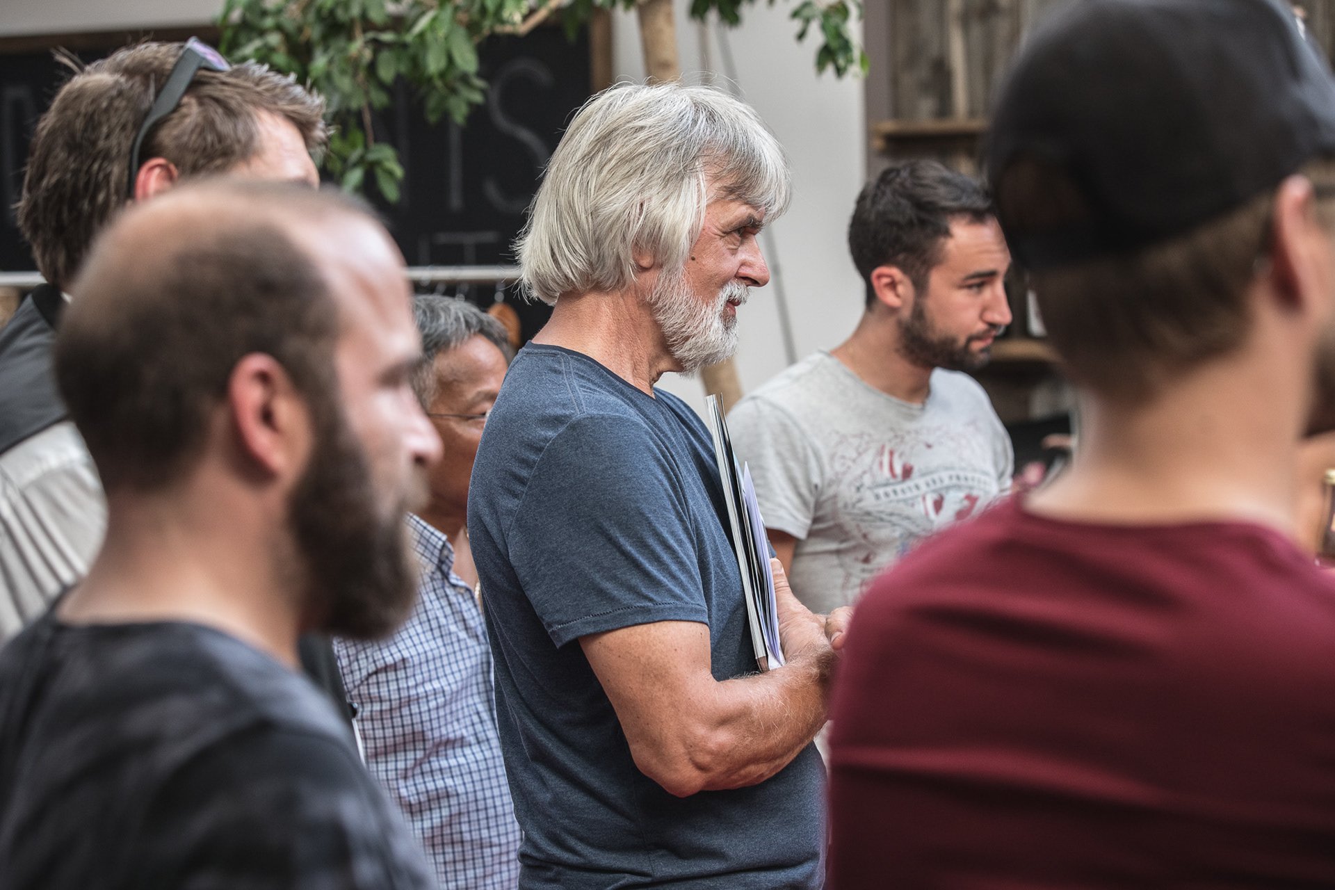 Eine Gruppe von Menschen, die in einem Raum zusammenstehen, darunter ein älterer Mann mit grauem Haar, der ein Notizbuch hält. Einige Menschen tragen lässige Kleidung und der Raum hat pflanzenartiges Dekor.