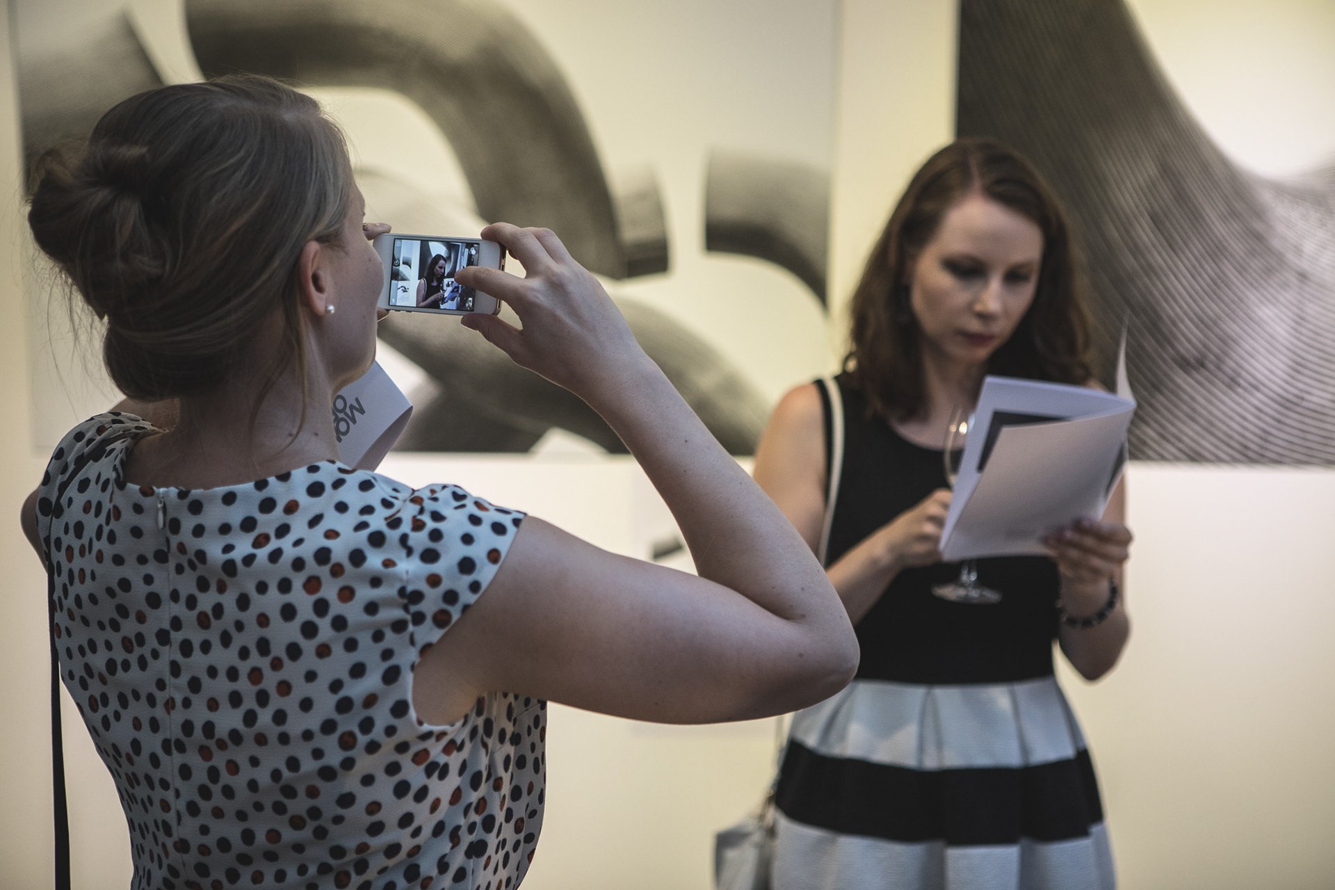 Zwei Frauen in einer Galerie; eine macht Fotos mit ihrem Handy, die andere liest ein Dokument; im Hintergrund abstrakte Kunst.