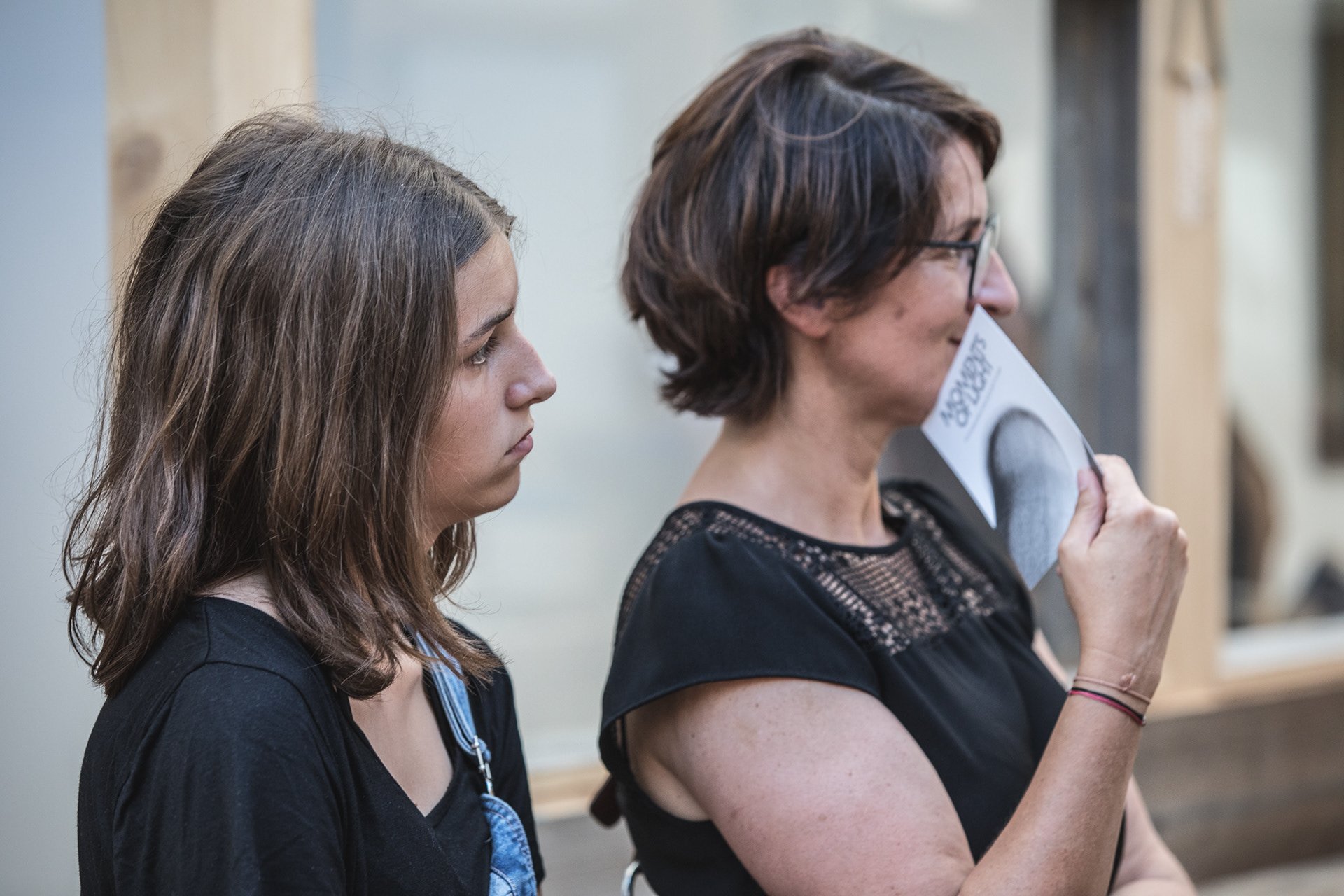 Zwei Frauen stehen nebeneinander. Eine Frau hält ein Foto oder Dokument vor ihr Gesicht. Es scheint ein informelles Treffen oder eine Veranstaltung zu sein. Beide Frauen tragen dunkle Kleidung.