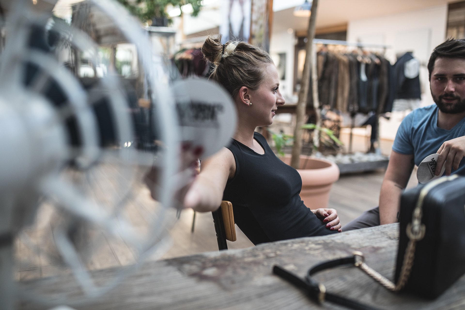Eine Frau sitzt an einem Tisch in einem Innenraum, mit einem Ventilator vor sich. Neben ihr sitzt ein Mann. Im Hintergrund sind Pflanzen und Kleidung auf einem Gestell zu sehen.