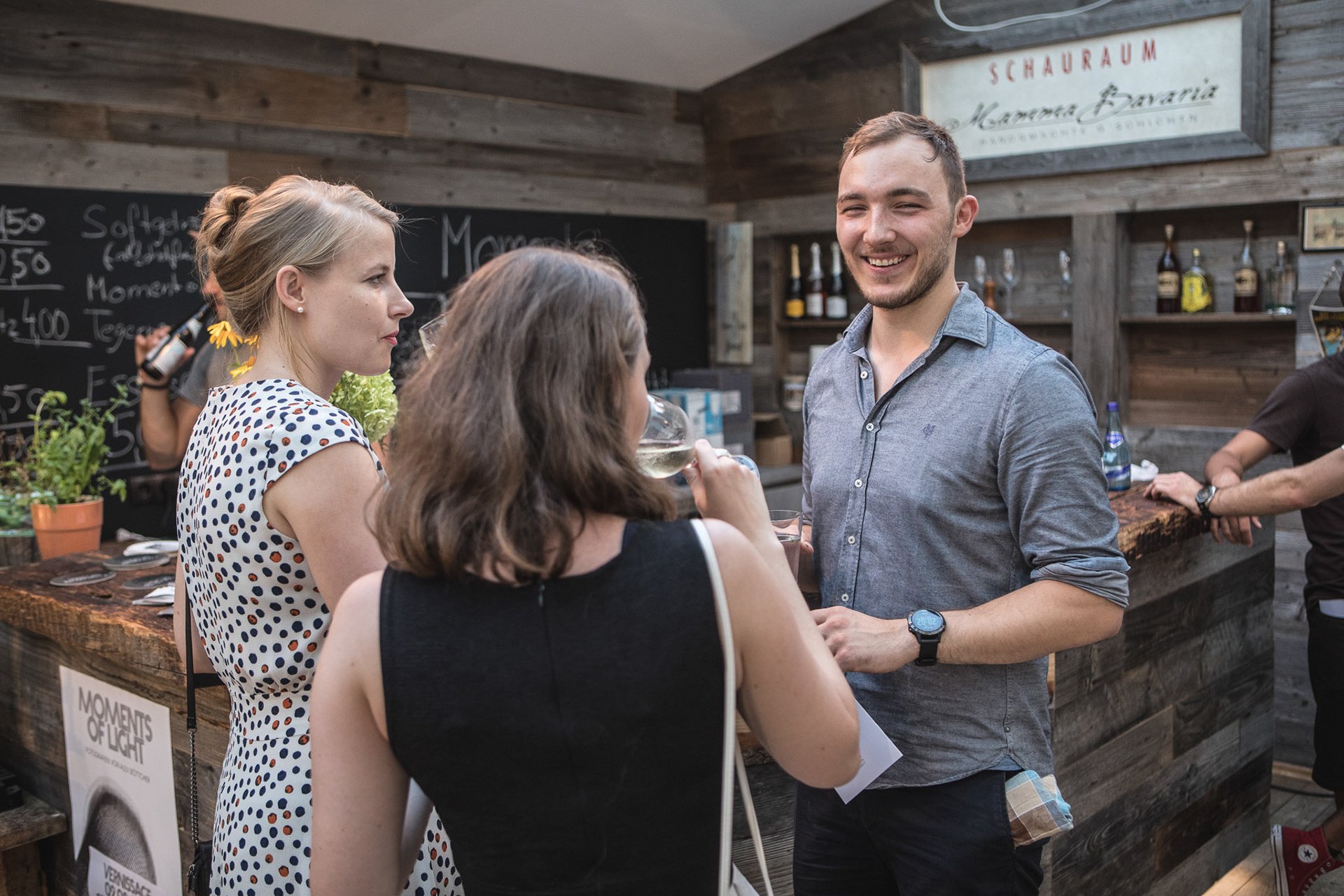 Gruppe von Menschen in einer Bar oder einem Café, die sich unterhalten. Eine Frau trägt ein gepunktetes Kleid, eine andere trinkt ein Glas Weißwein. Im Hintergrund ist eine Bar mit Getränken zu sehen.