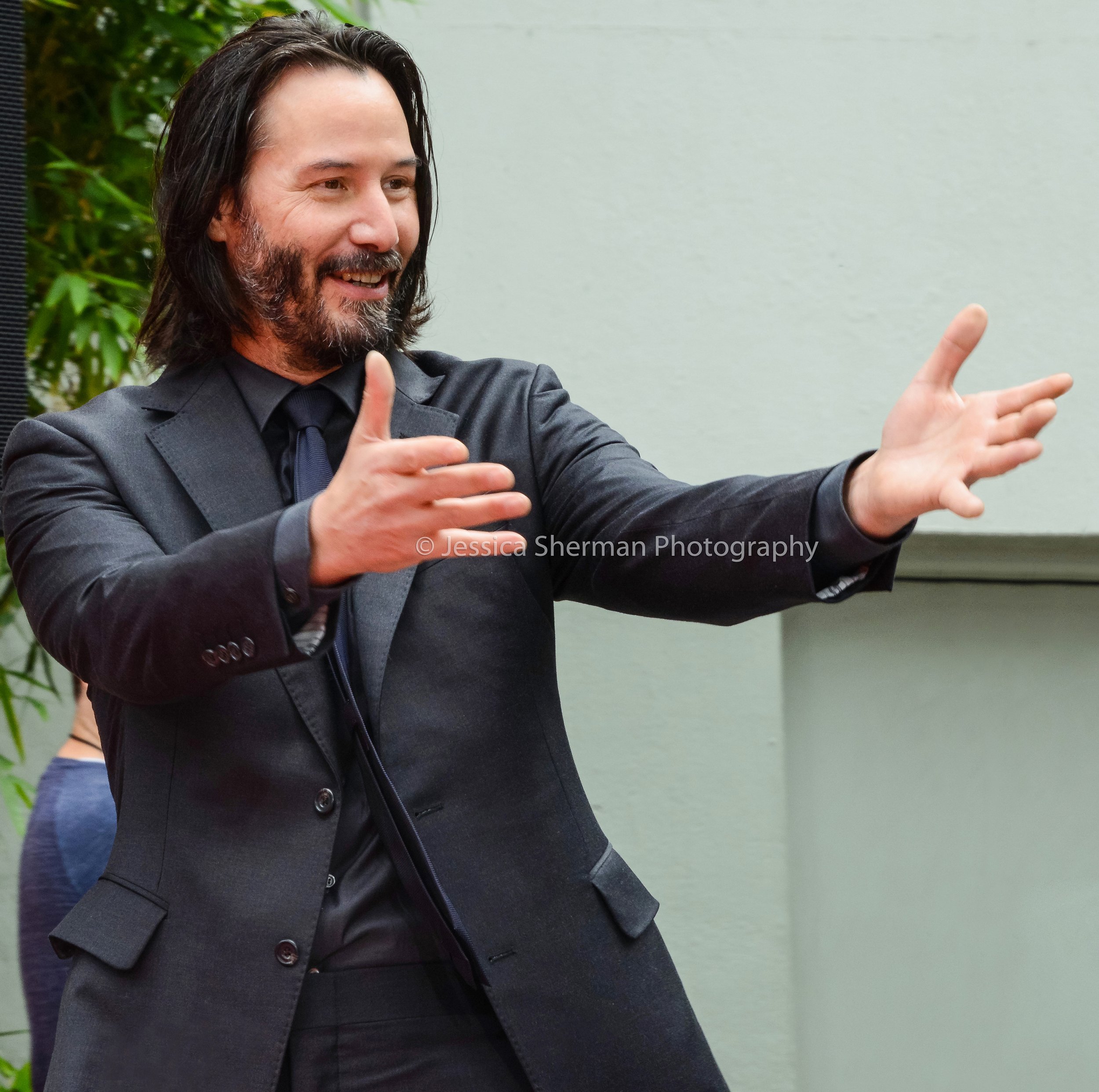 Keanu Reeves in a black suit and black tie with long dark hair and a beard, smiling and gesturing with his hands as if explaining something.
