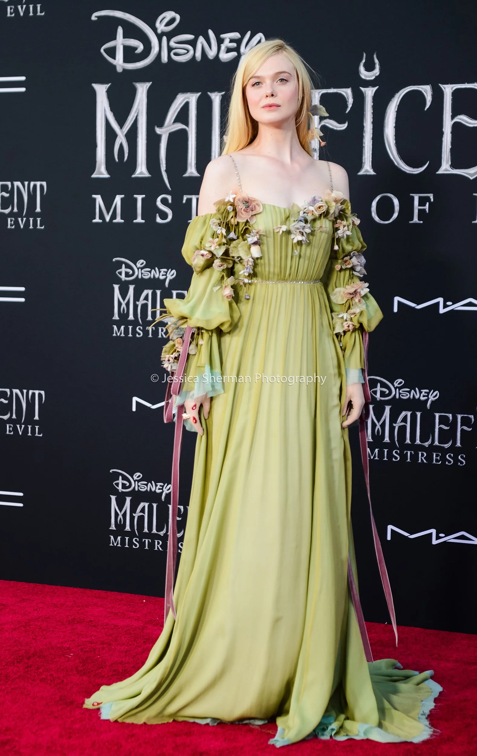 A woman in a long, flowing yellow-green gown with floral embellishments on the sleeves and bodice, standing on a red carpet at a Disney "Maleficent" movie event.