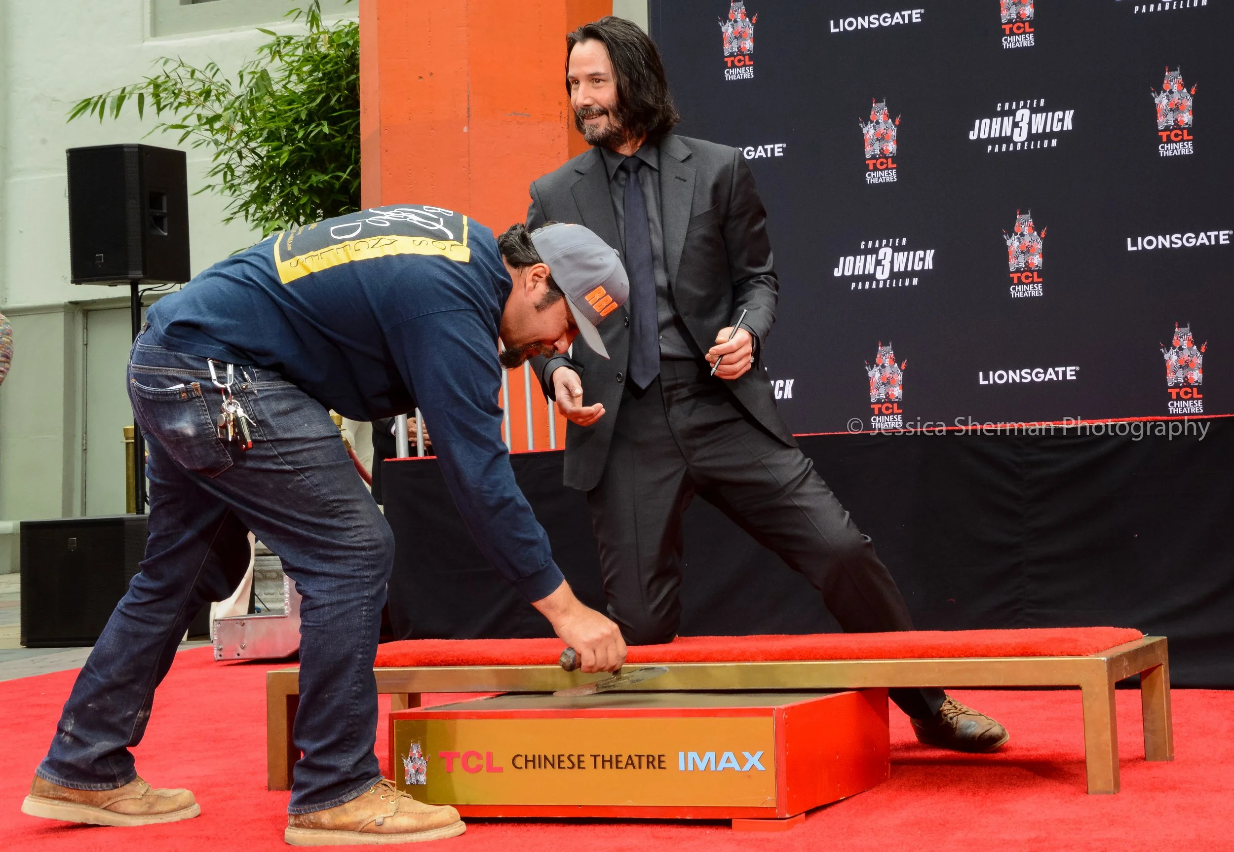 A man in a dark suit with long hair and a beard is standing on a red carpeted stage, holding a pen. Another man in casual clothing, including a blue hoodie, jeans, and a gray cap, is bending down and hammering a nail into a wooden frame. The backdrop