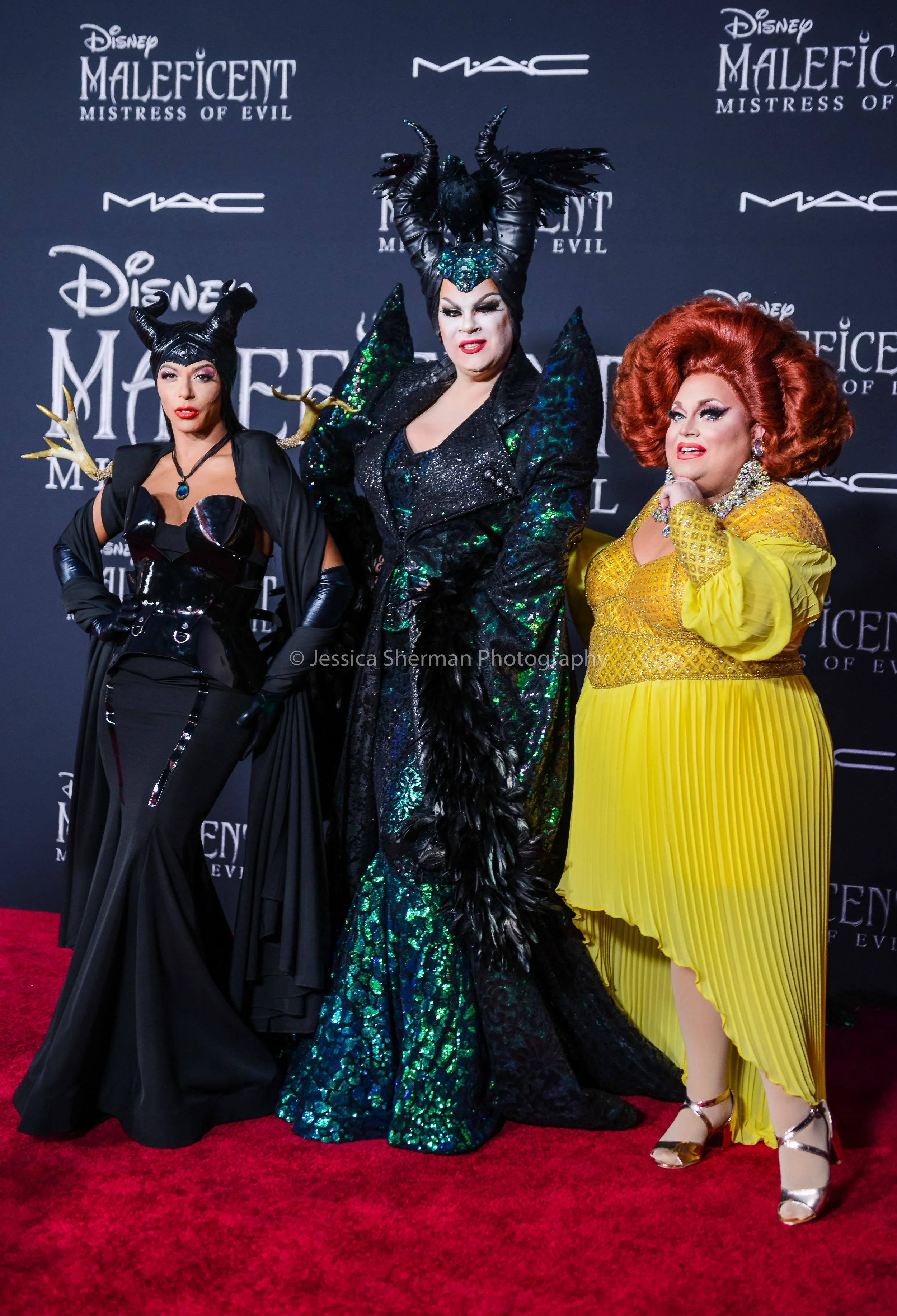 Three women dressed as characters from Disney's Maleficent movie standing on a red carpet with a black backdrop featuring logos of Disney, MAC, and Maleficent Mistress of Evil.