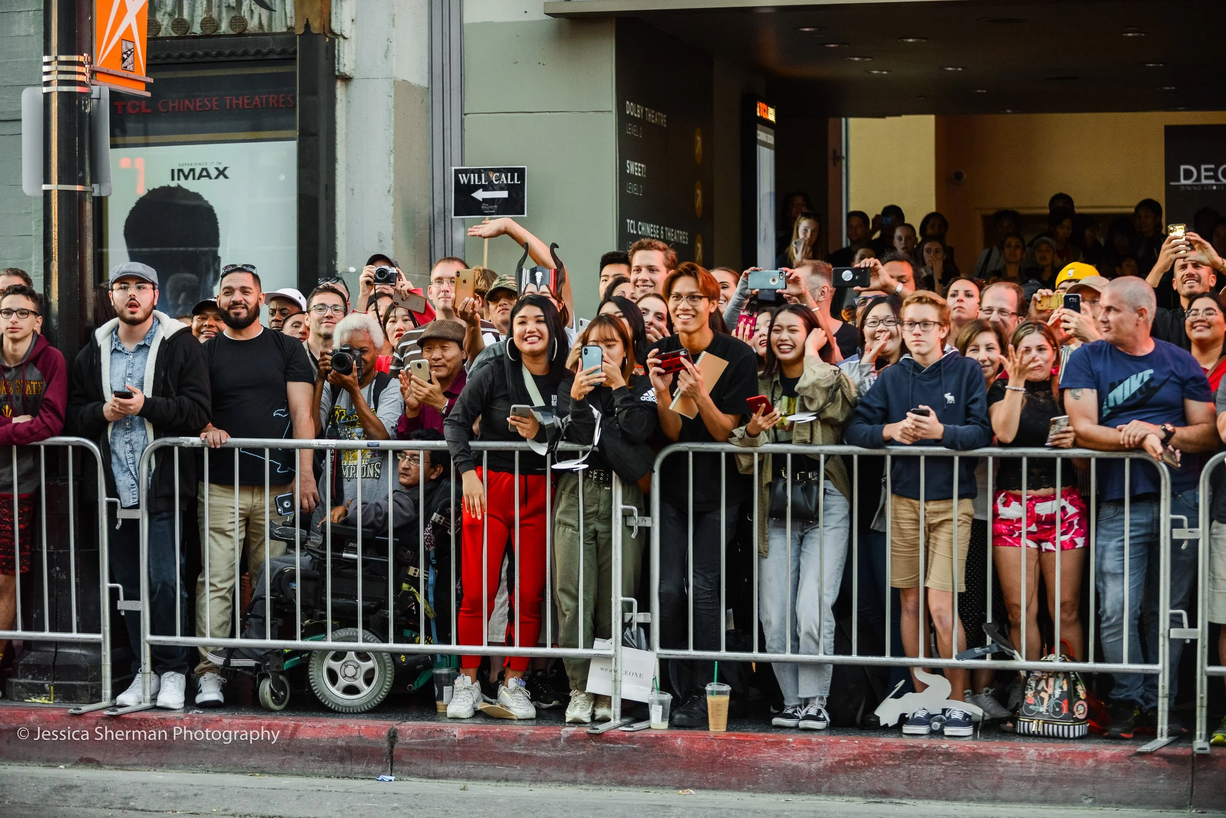 Crowd of people behind metal barricades, smiling and taking photos during a public event, with some holding phones and cameras. The background shows a theater entrance and signs.