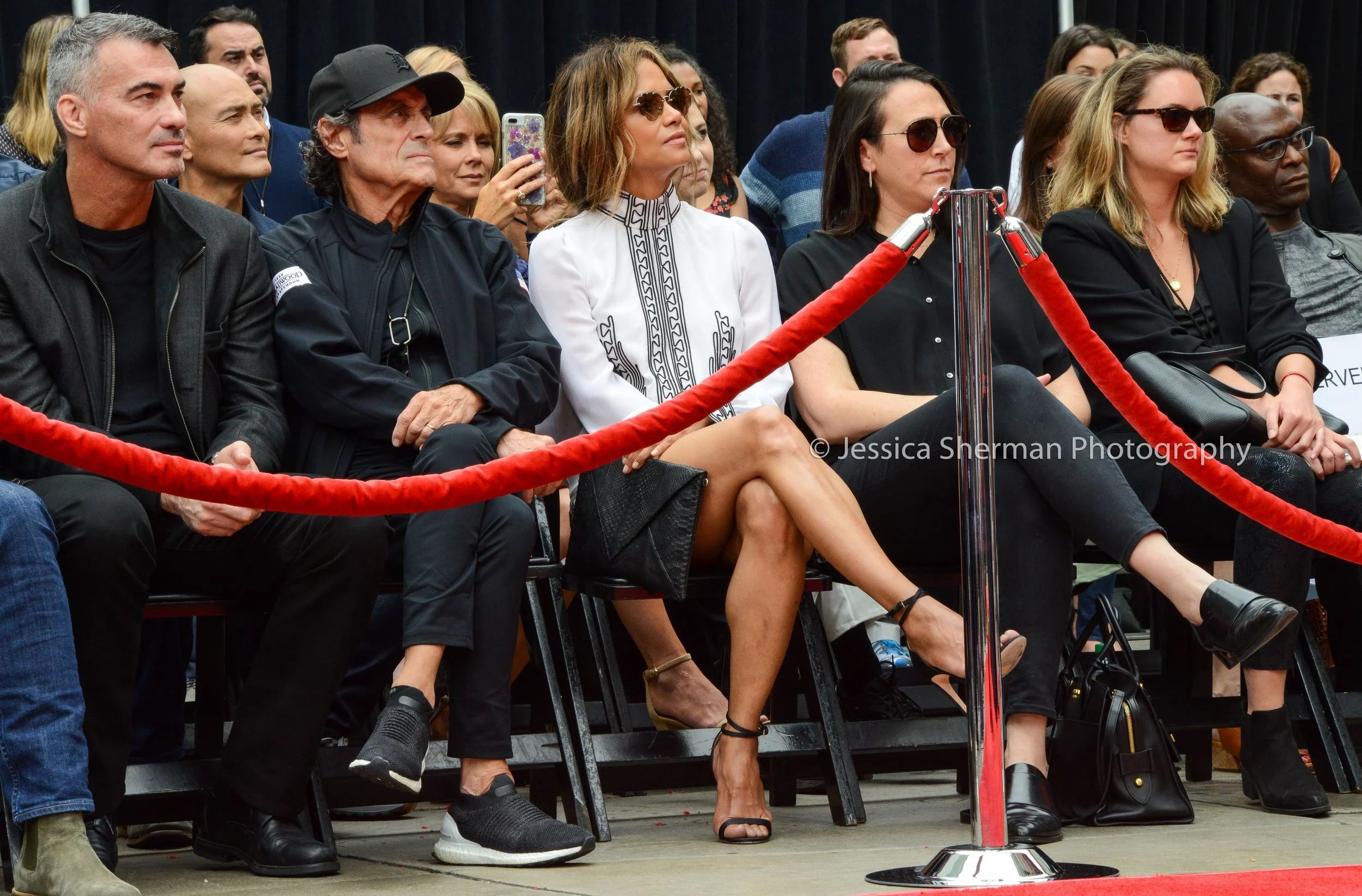 People sitting behind a red velvet rope at an event, dressed in black, white, and casual attire, with some wearing sunglasses, during daytime.