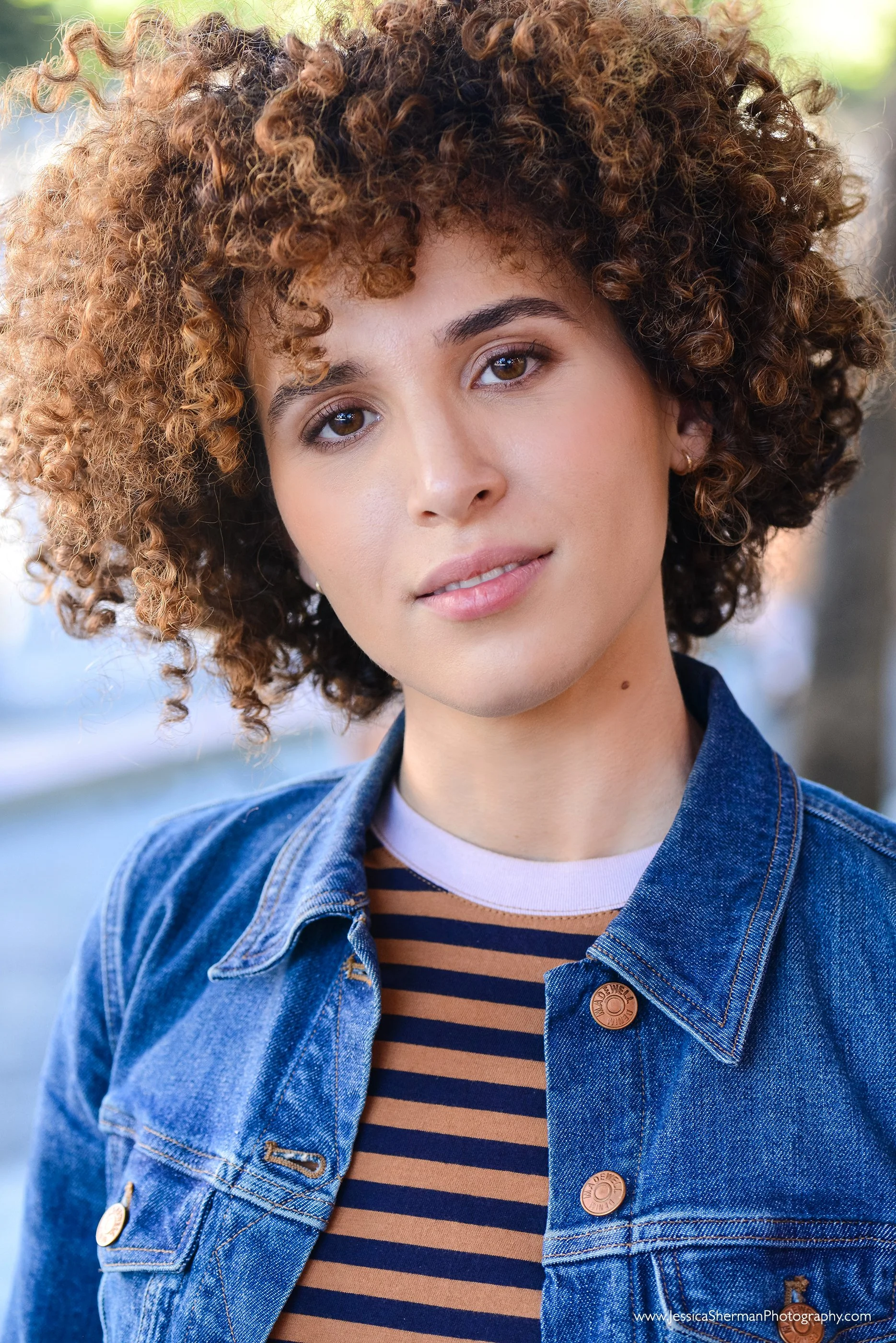 A young woman with curly brown hair, light skin, and brown eyes, wearing a denim jacket over a brown and navy striped shirt, outdoors on a sunny day.