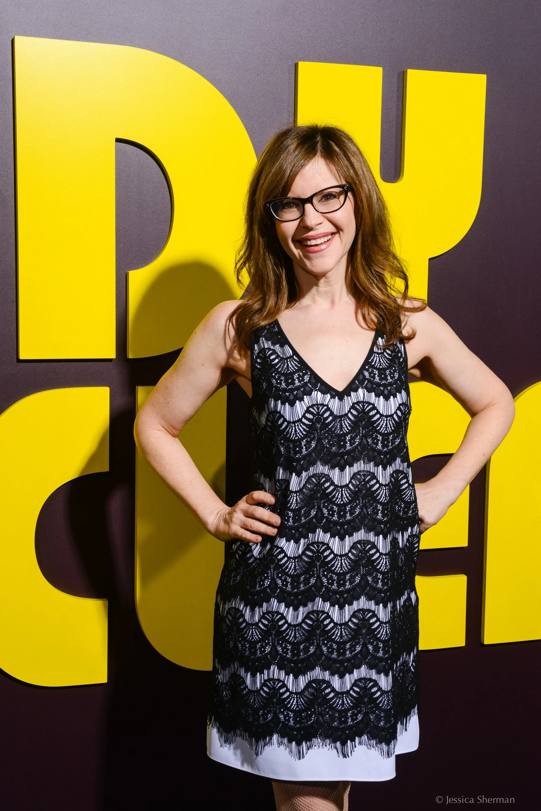 A woman with long red hair and glasses smiling, standing in front of large yellow letters on a purple background, wearing a black lace dress with a white lining.