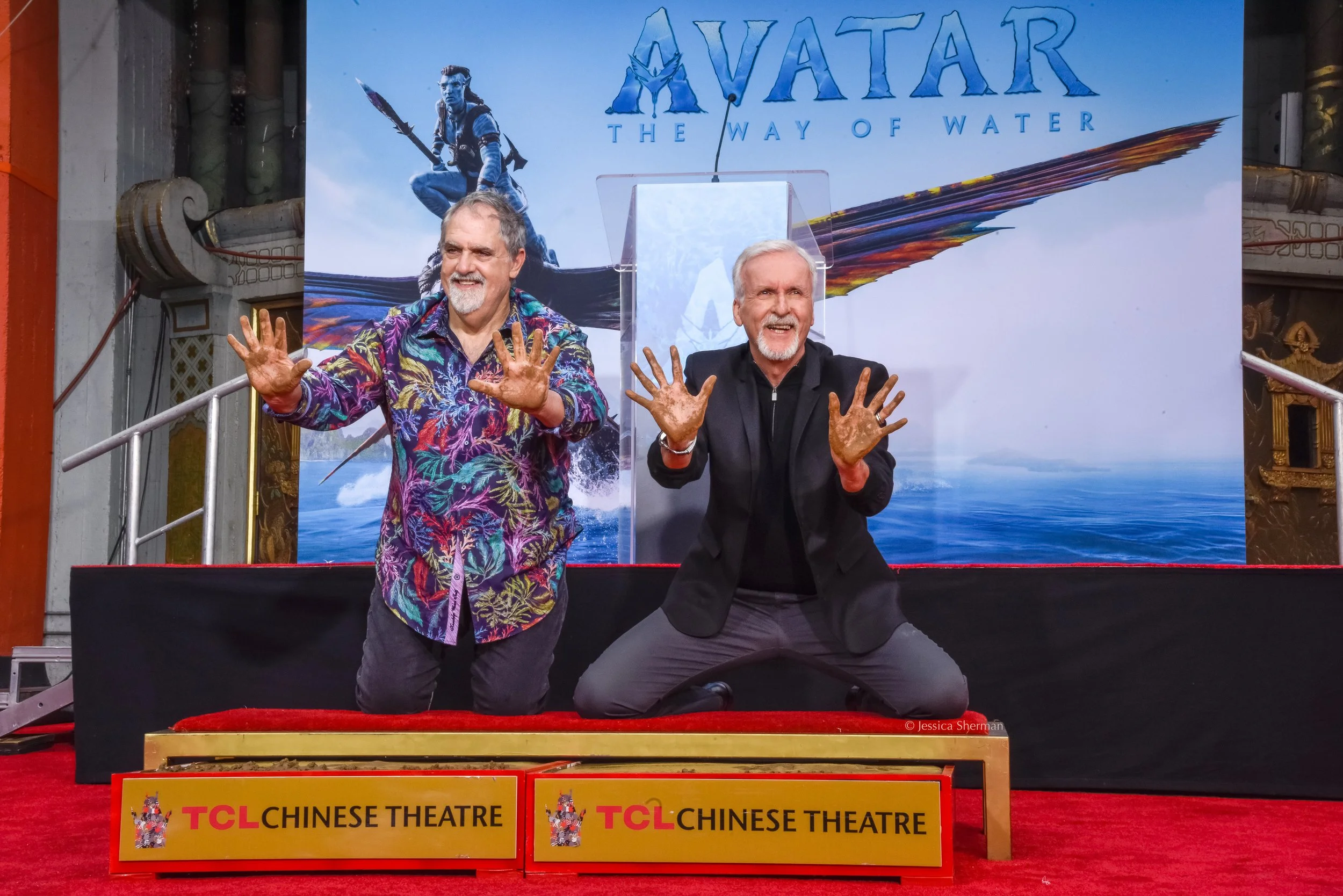 Two men kneeling on a stage with a backdrop of the movie 'Avatar: The Way of Water.' They are smiling and showing their hands, covered in what appears to be mud or dirt. There are signs in front of the stage that read 'TCL Chinese Theatre.'