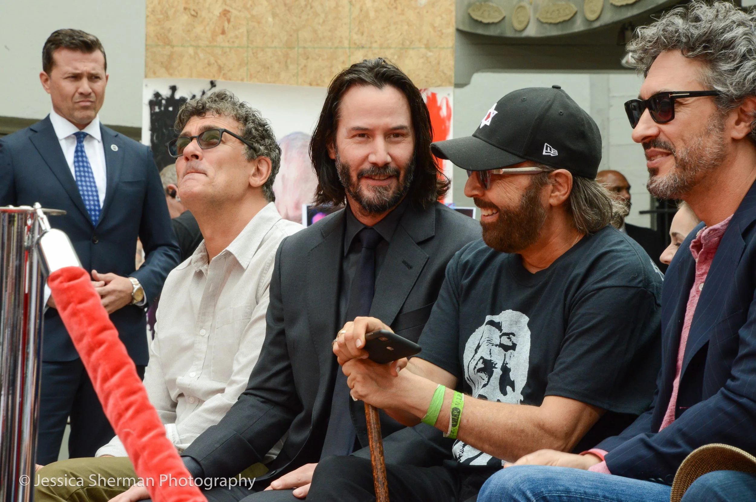 Keanu Reeves and friends seated in a row at an event, engaged in conversation, with one holding a smartphone, dressed in casual and formal clothing, and a security guard standing in the background.